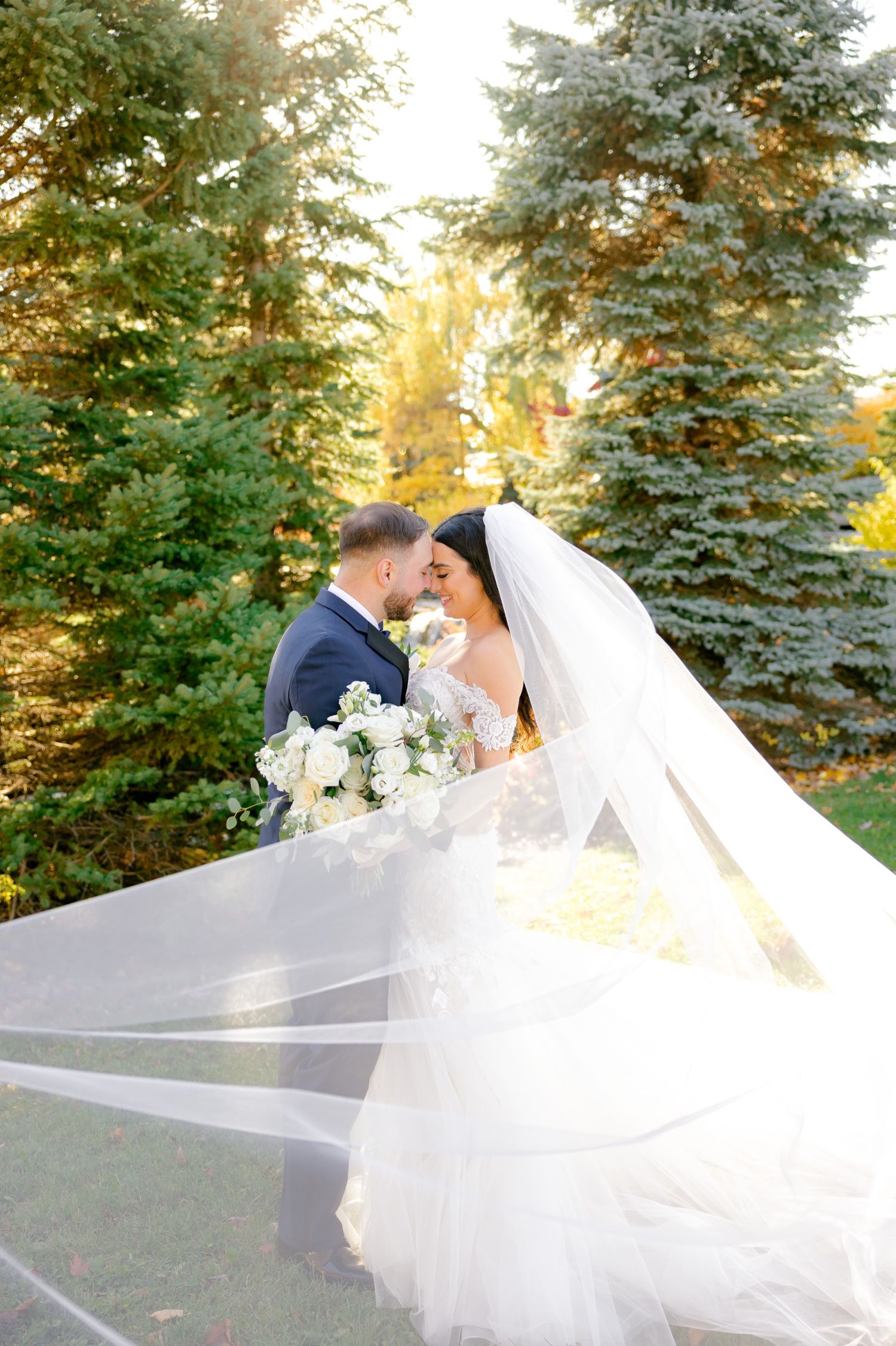 Wedding couple poses among evergreen trees with flowing white veil in autumn light.