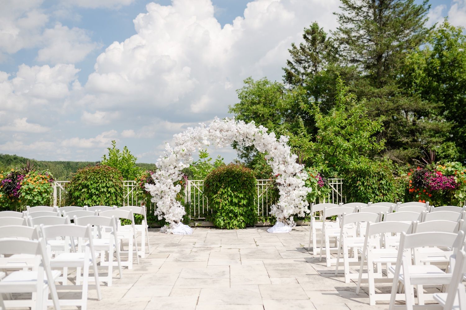 Romantic outdoor wedding ceremony setup with white floral arch and chairs on stone patio.
