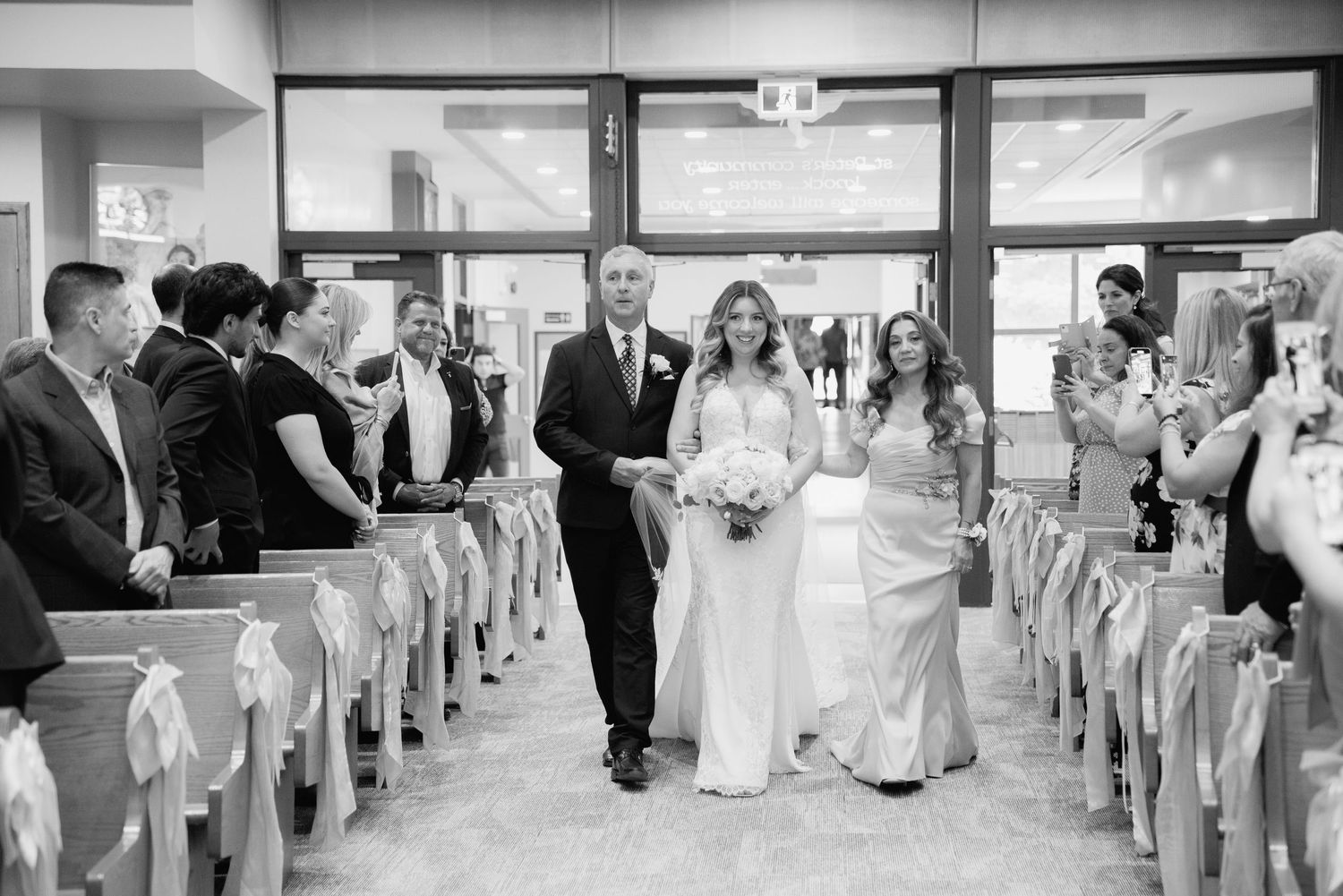Black and white photo of wedding processional walking down church aisle lined with wooden pews.