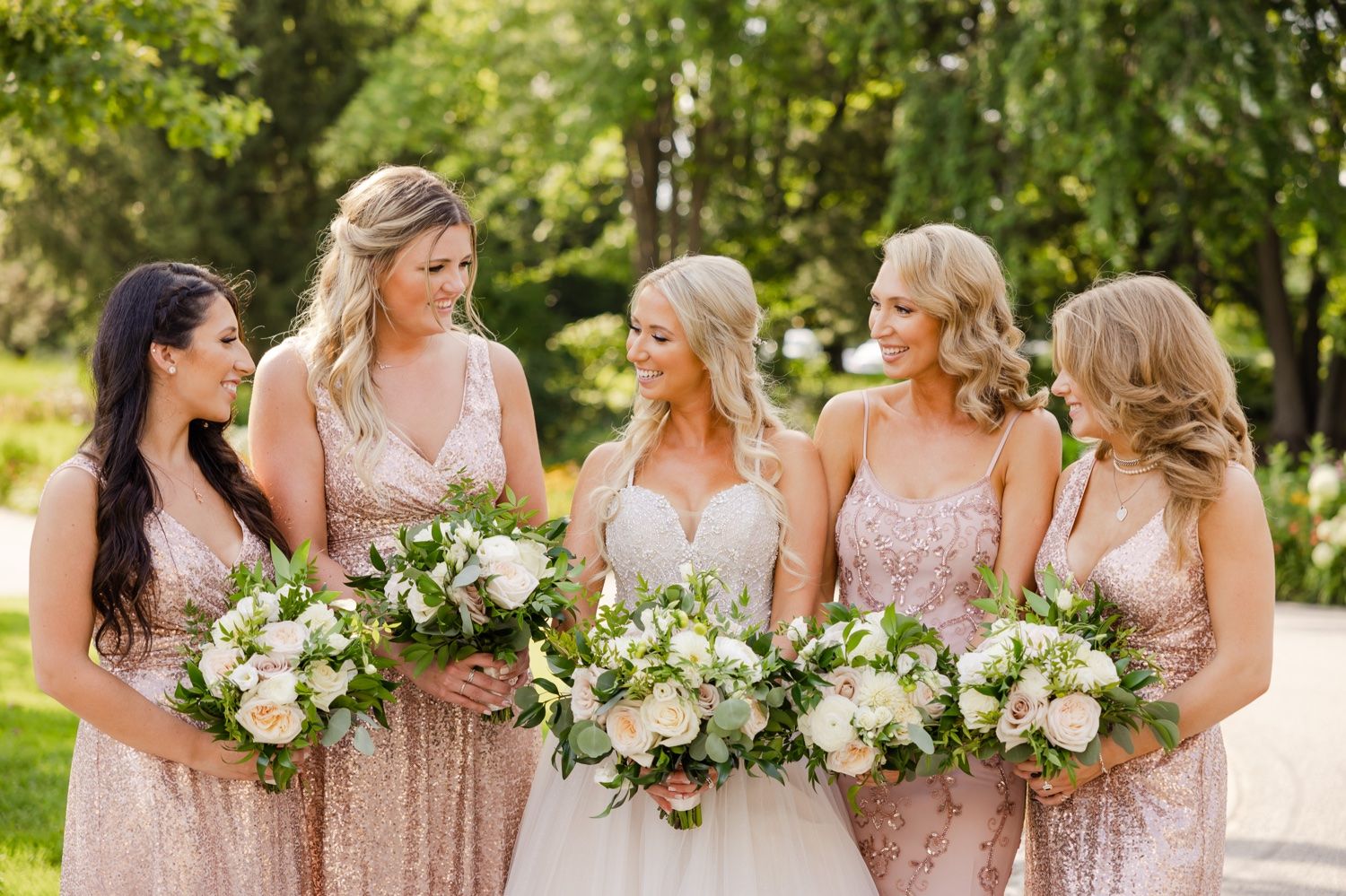 Bridal party in sparkling pink gowns holding elegant white and green bouquets in an outdoor setting.