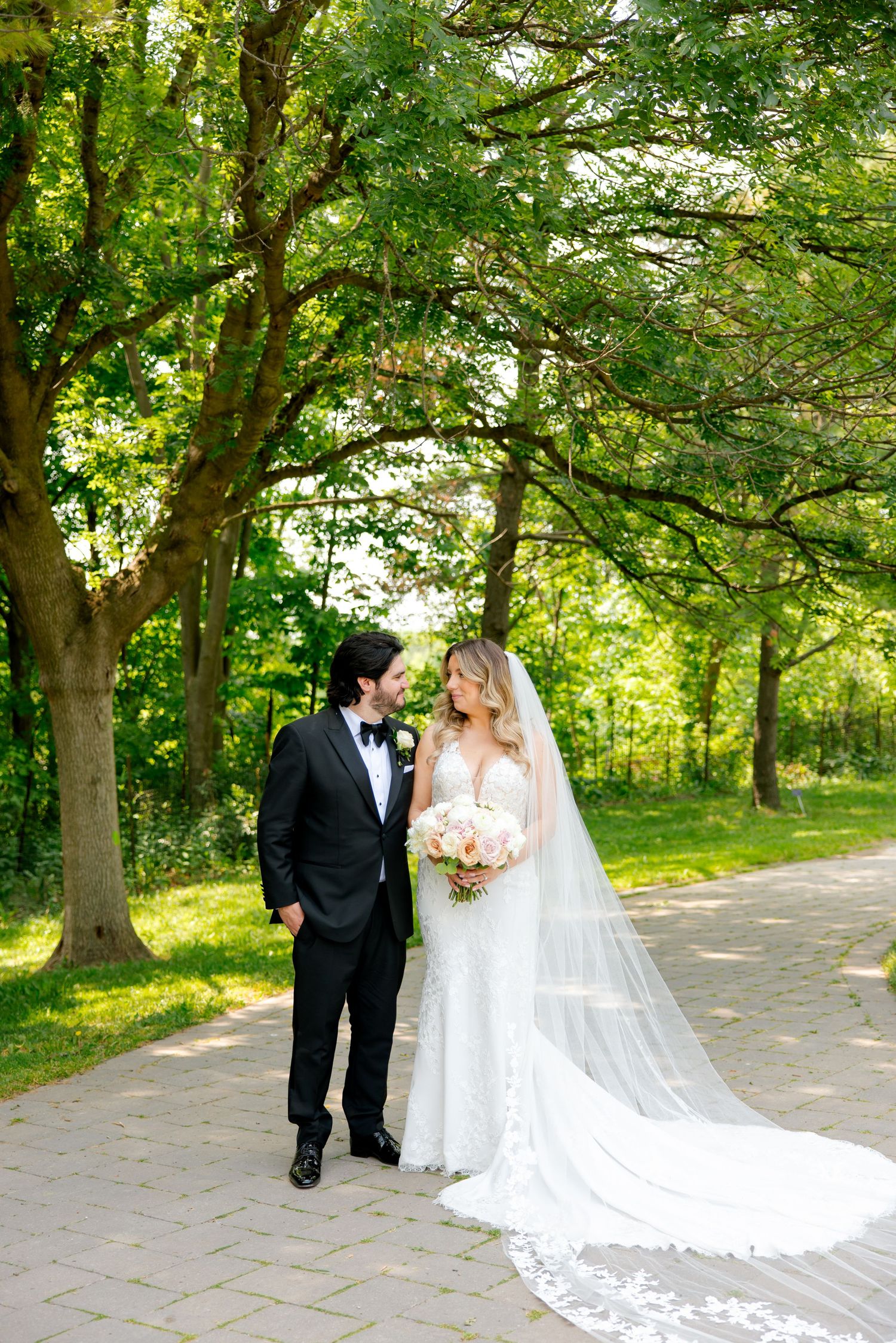 A newlywed couple stands on a scenic garden path surrounded by lush green trees.