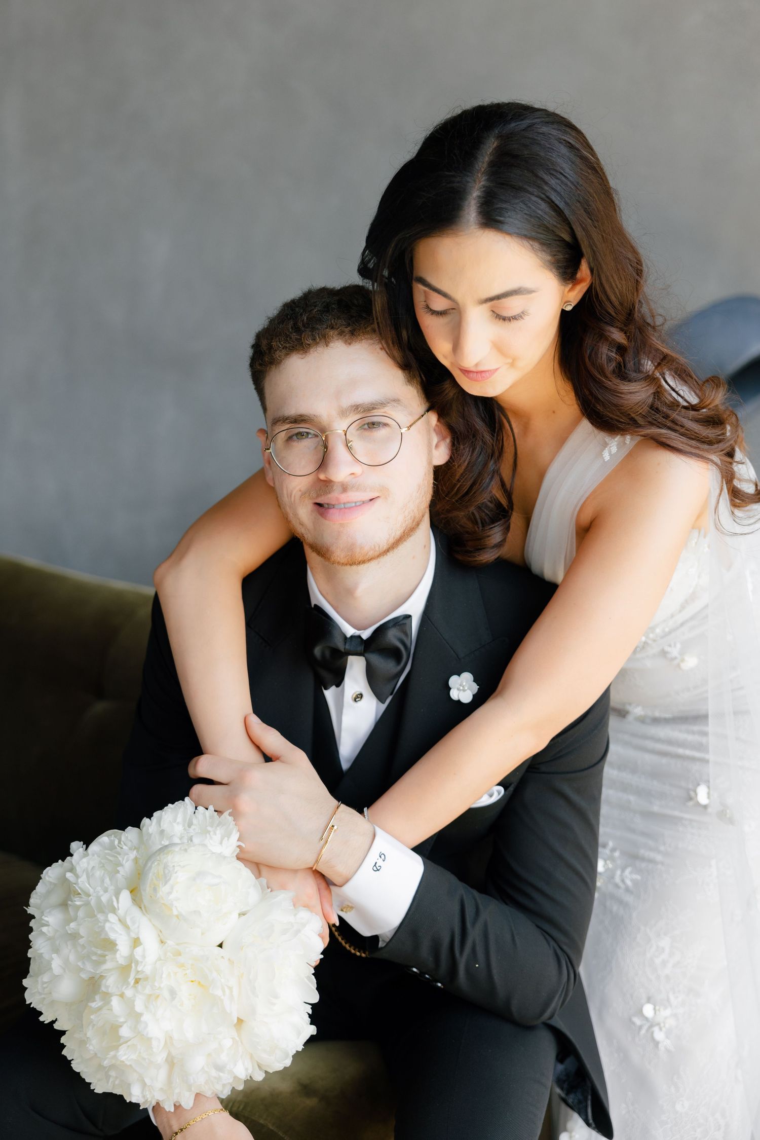 Intimate wedding portrait of couple embracing on dark couch with white floral bouquet.