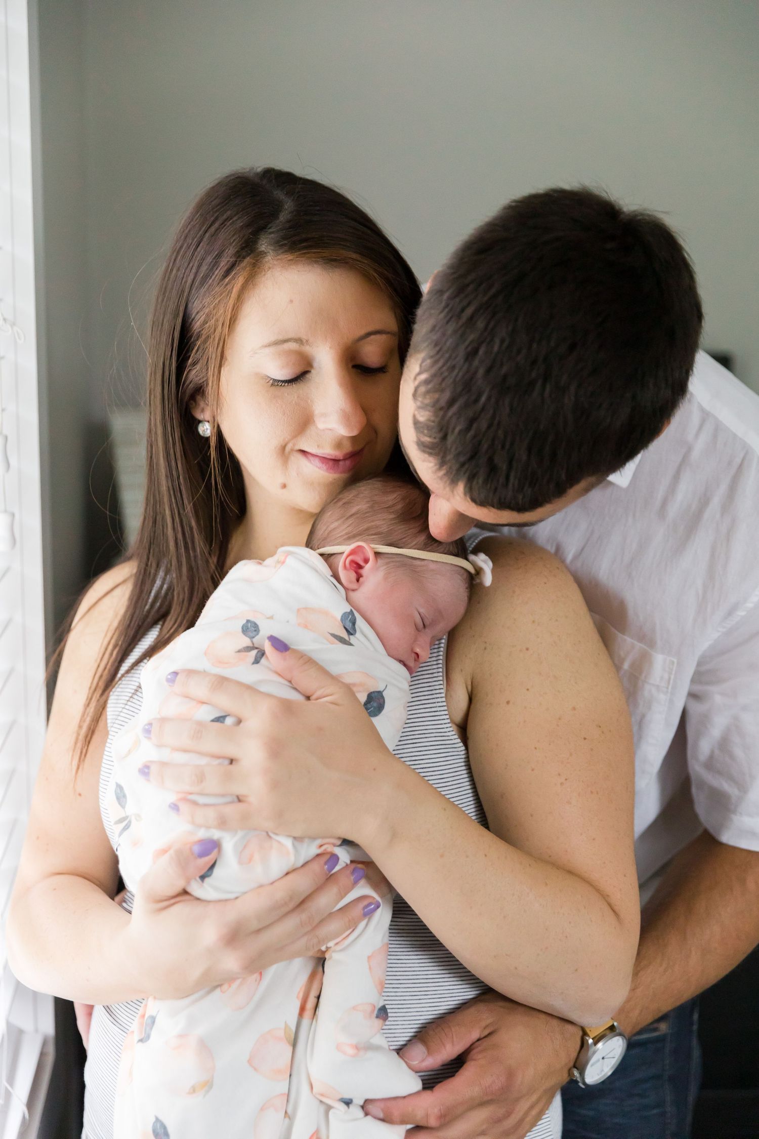 Parents sharing intimate moment with newborn by window.