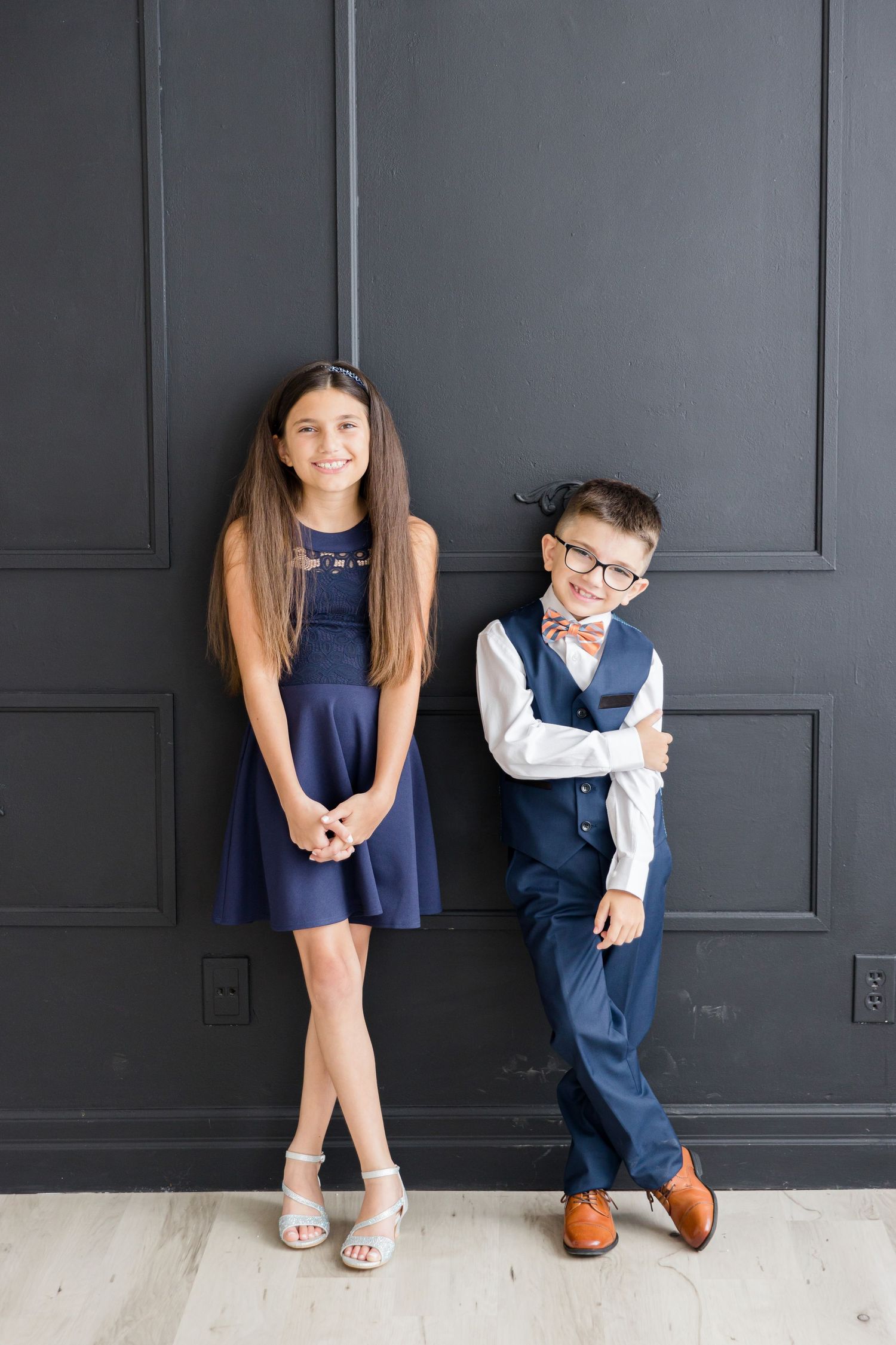 Two children in formal navy attire pose against a dark gray wall with classic architectural details.