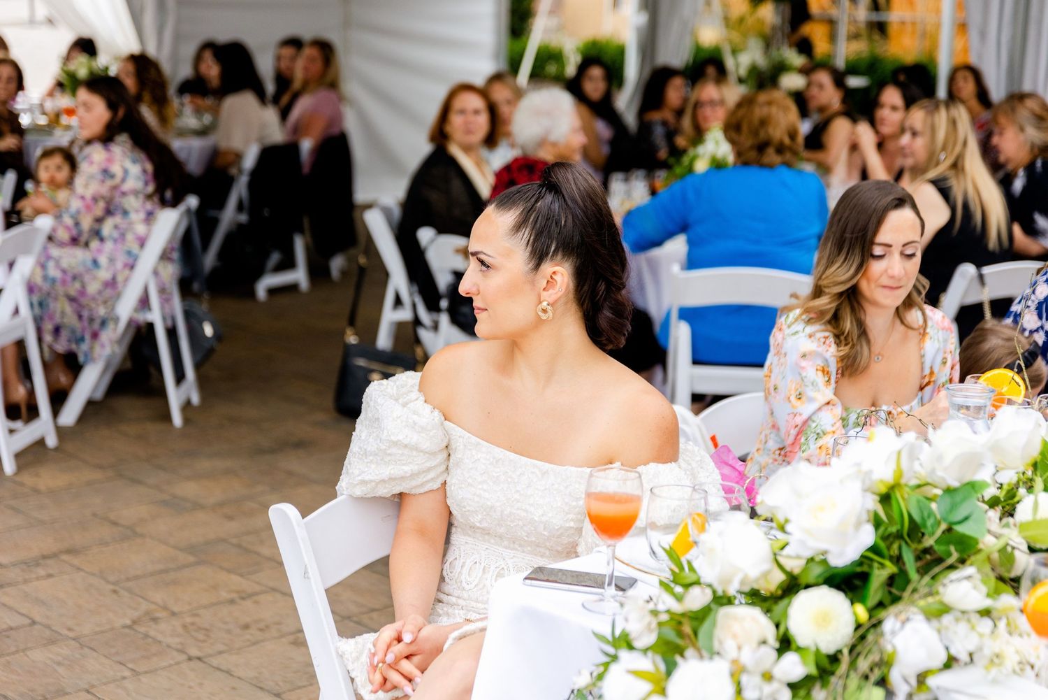Wedding celebration in an outdoor tent venue with white chairs and elegant floral arrangements.