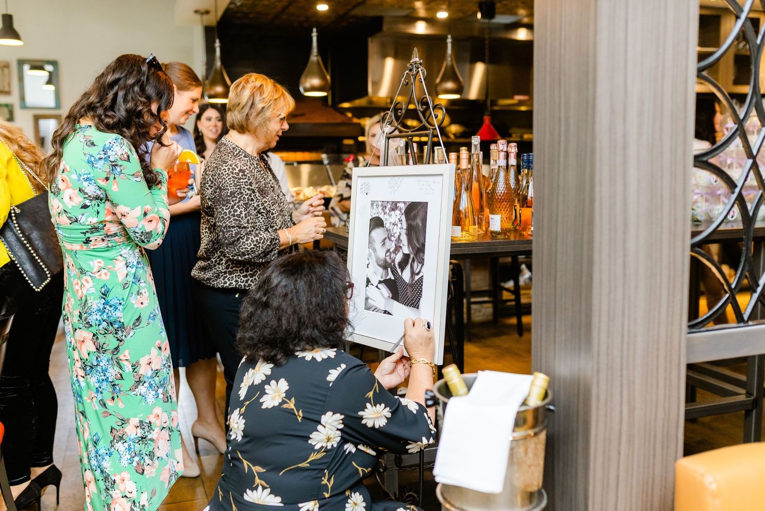 Guests viewing frames and decorations at an elegant indoor event space with wine bottles on display.
