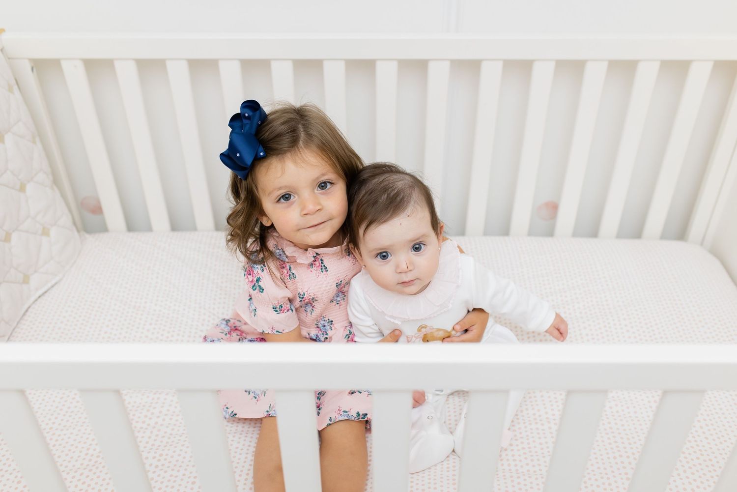 Two children pose together in a white crib, one wearing pink and blue.