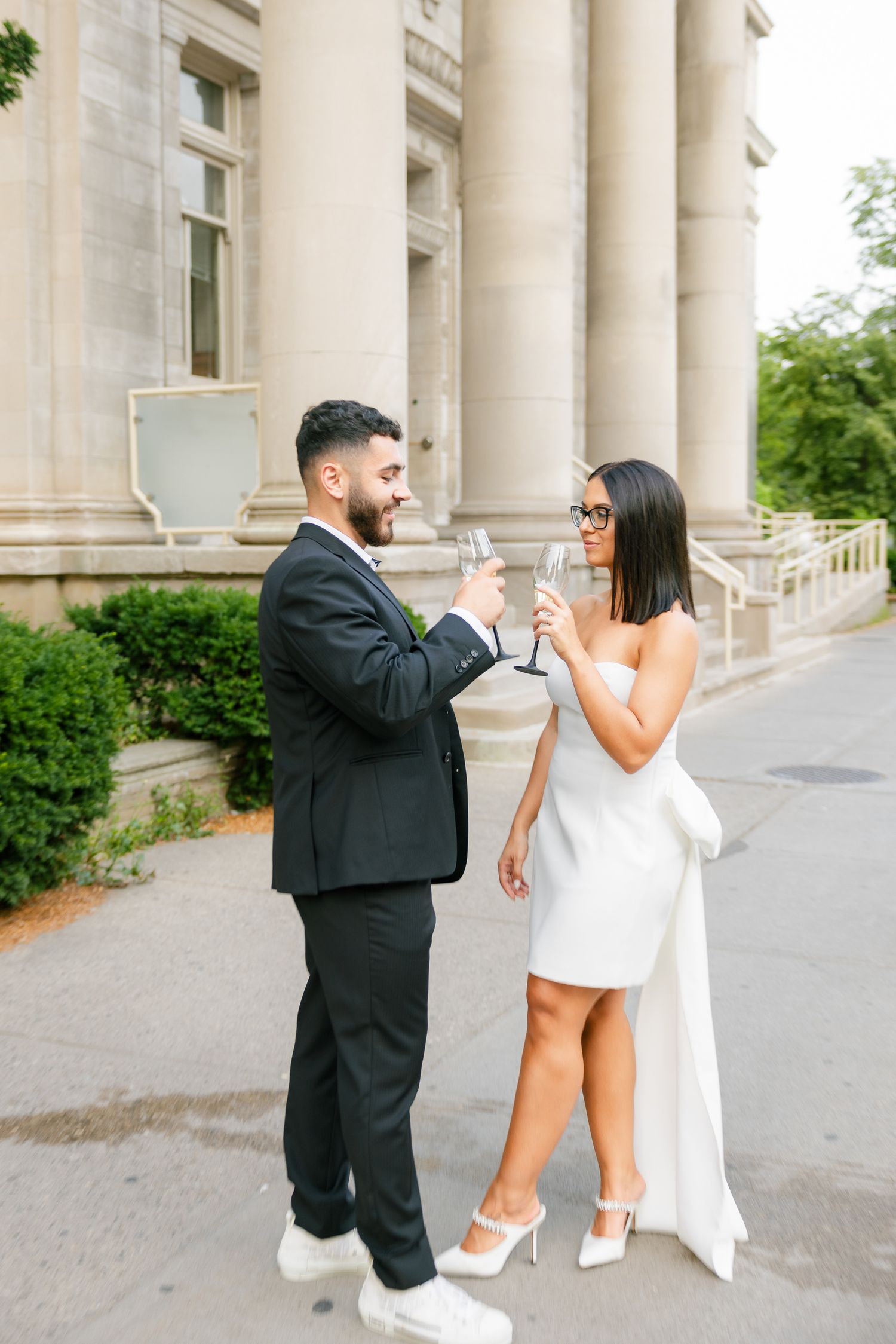 A couple in formal attire laughs and toasts with champagne glasses outside a columned building.
