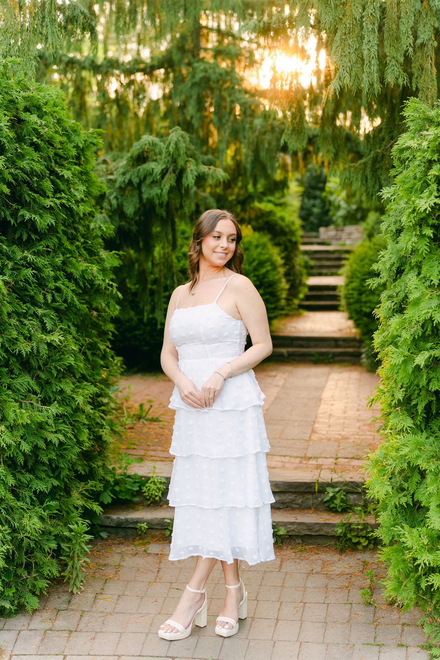A tiered white sundress worn in a lush garden path with willow trees and stone steps.