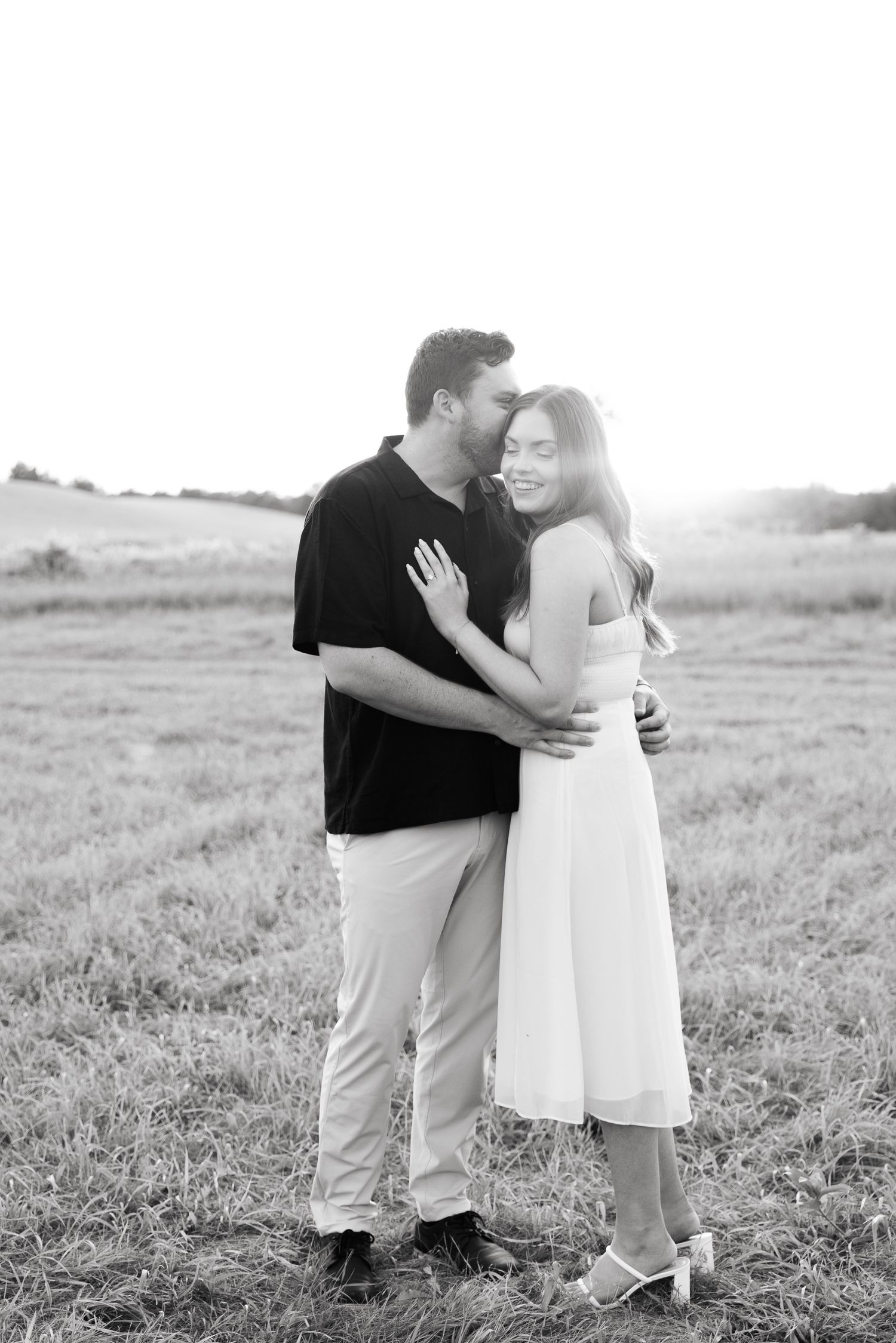 Romantic couple embraces in a serene field during sunset photoshoot in black and white.