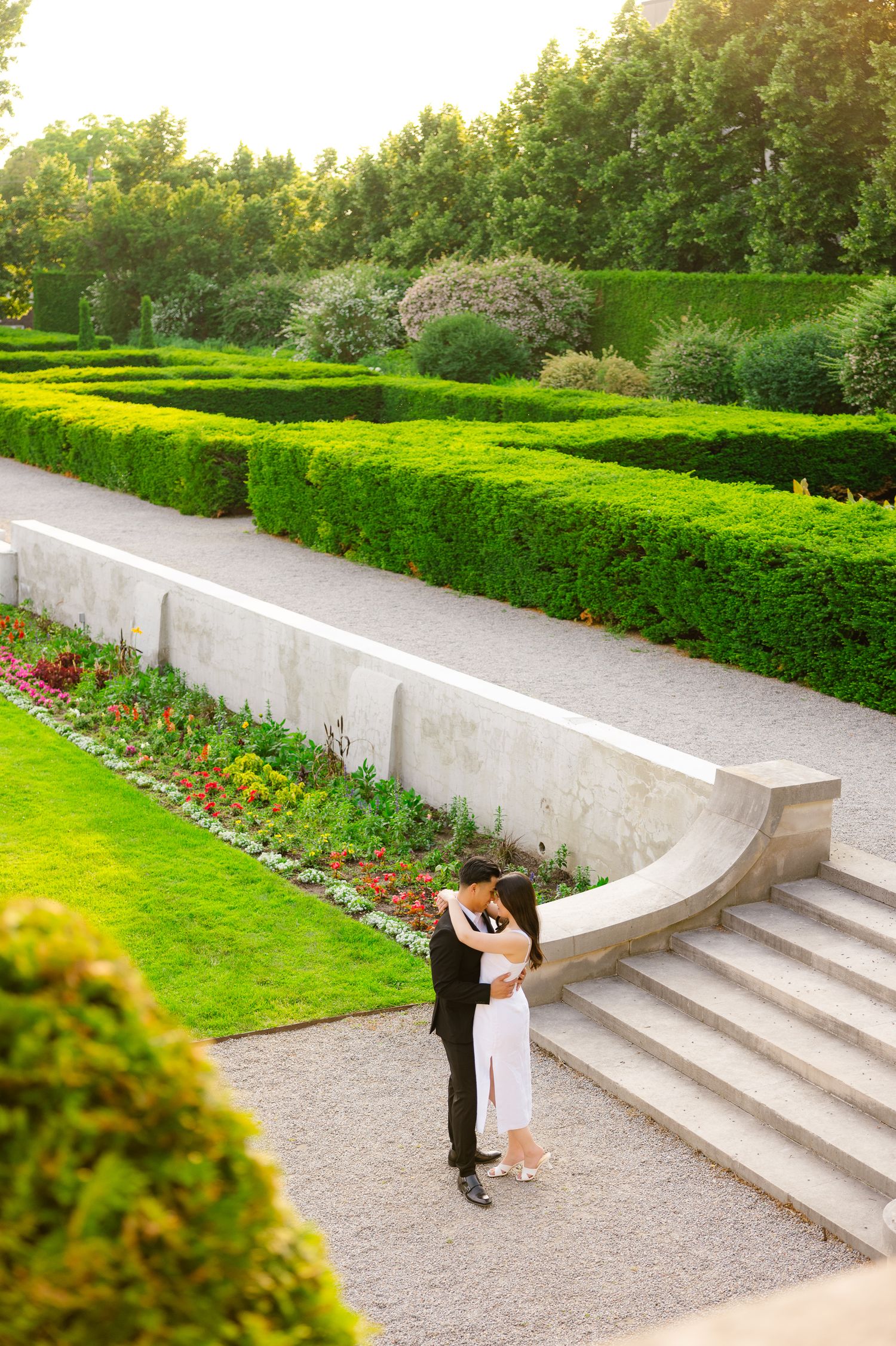 Sunlit garden scene with two people sharing a romantic moment on elegant stone steps.