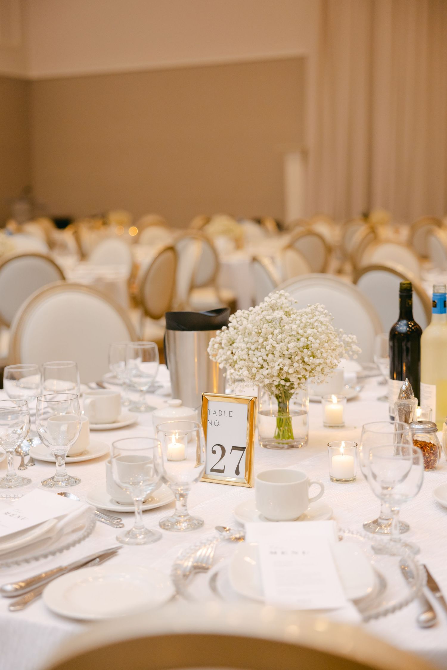 Wedding reception table setting with baby's breath centerpiece, table number, and cream-colored chairs.
