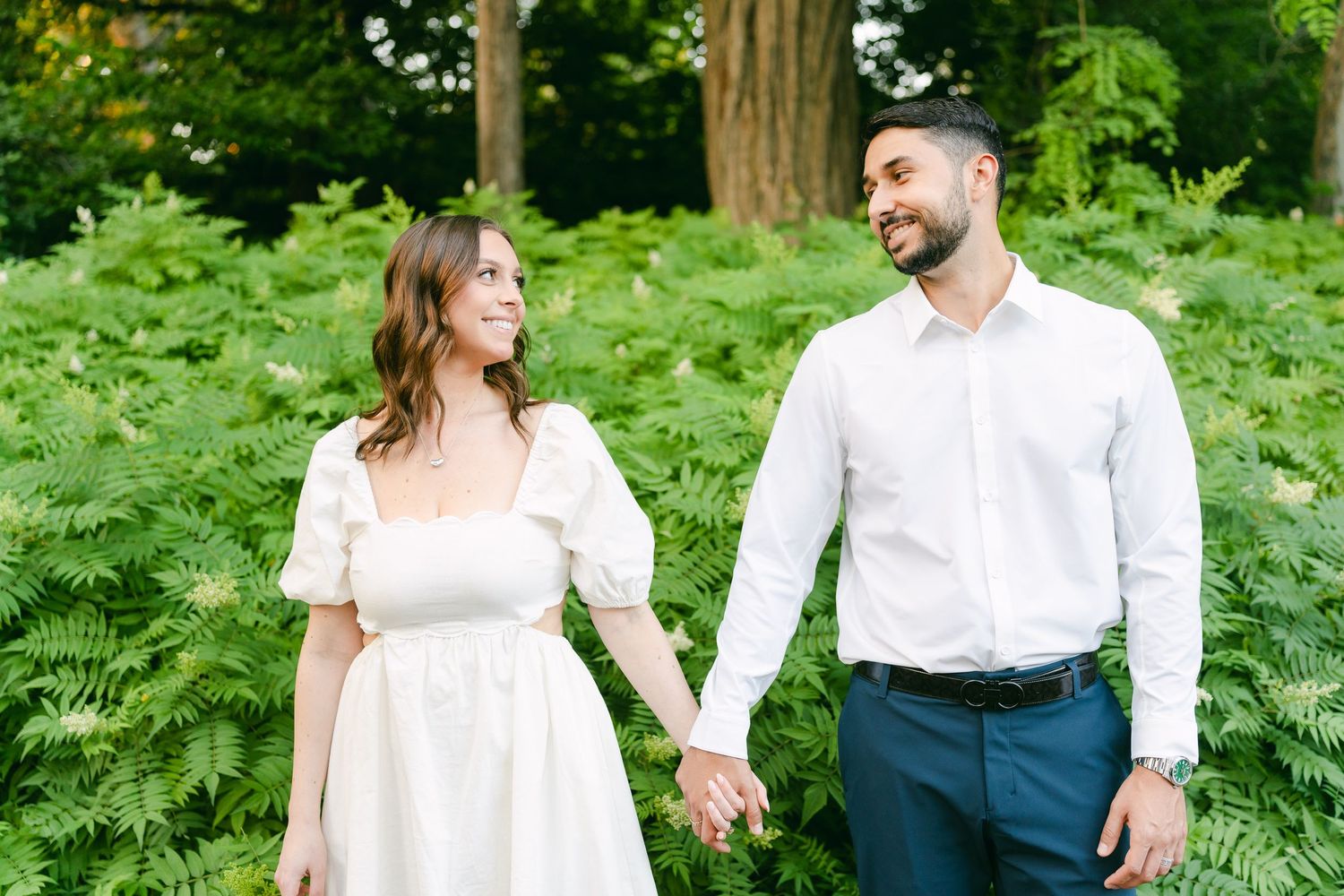 Couple holding hands while walking through fern-filled garden setting.