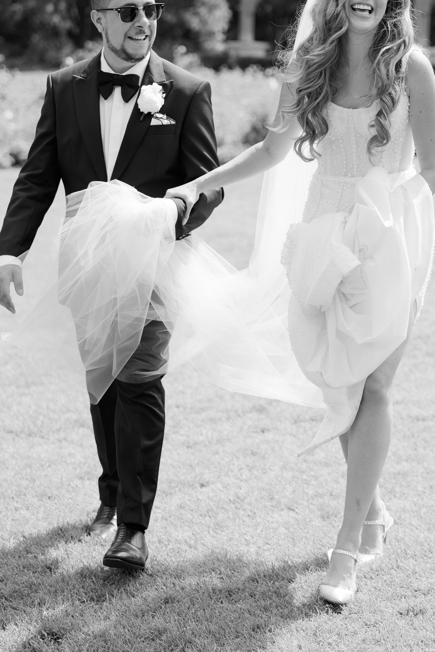 Black and white photo of a newlywed couple running playfully on grass with the bride's dress flowing.