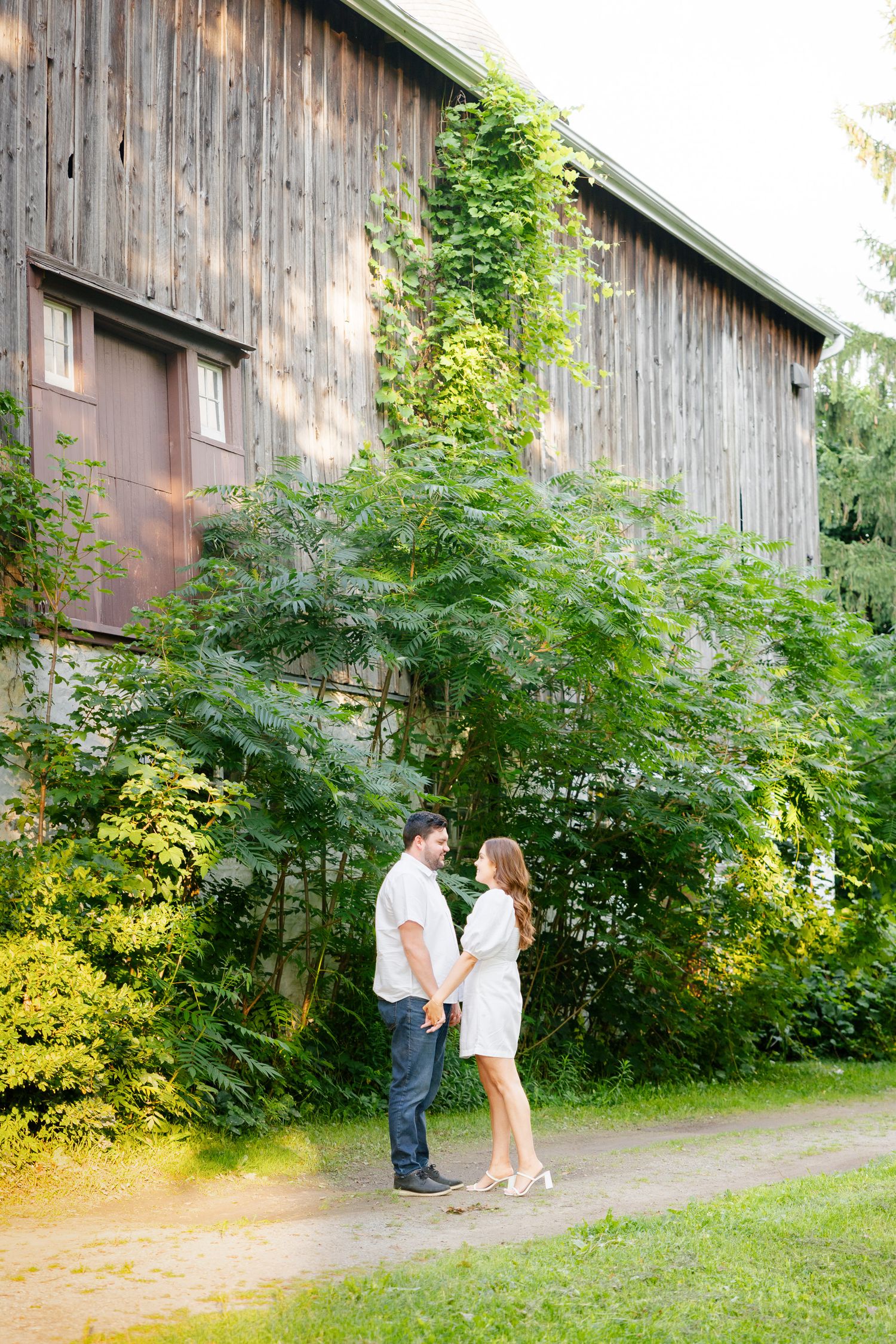 A couple stands intimately in front of a rustic wooden barn surrounded by greenery.