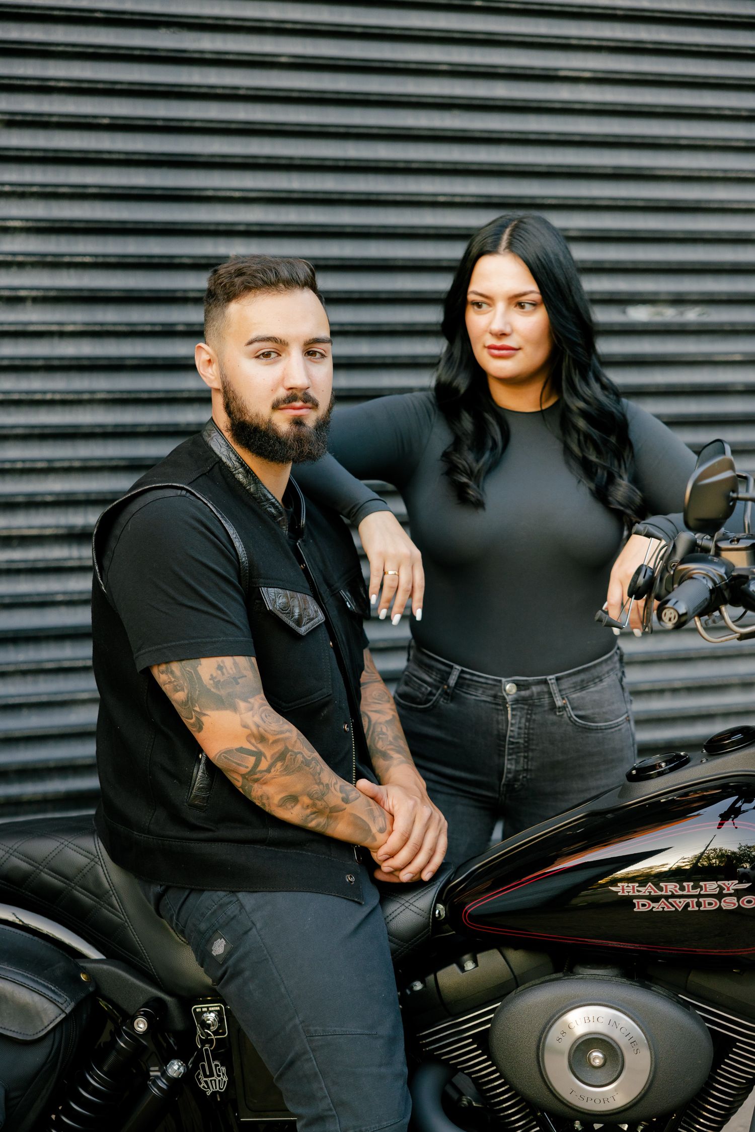 A couple in matching dark outfits poses confidently with their motorcycle.
