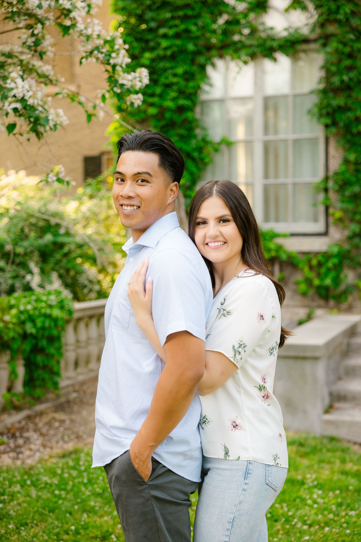 Couple in casual attire poses against ivy-covered building and flowering trees.