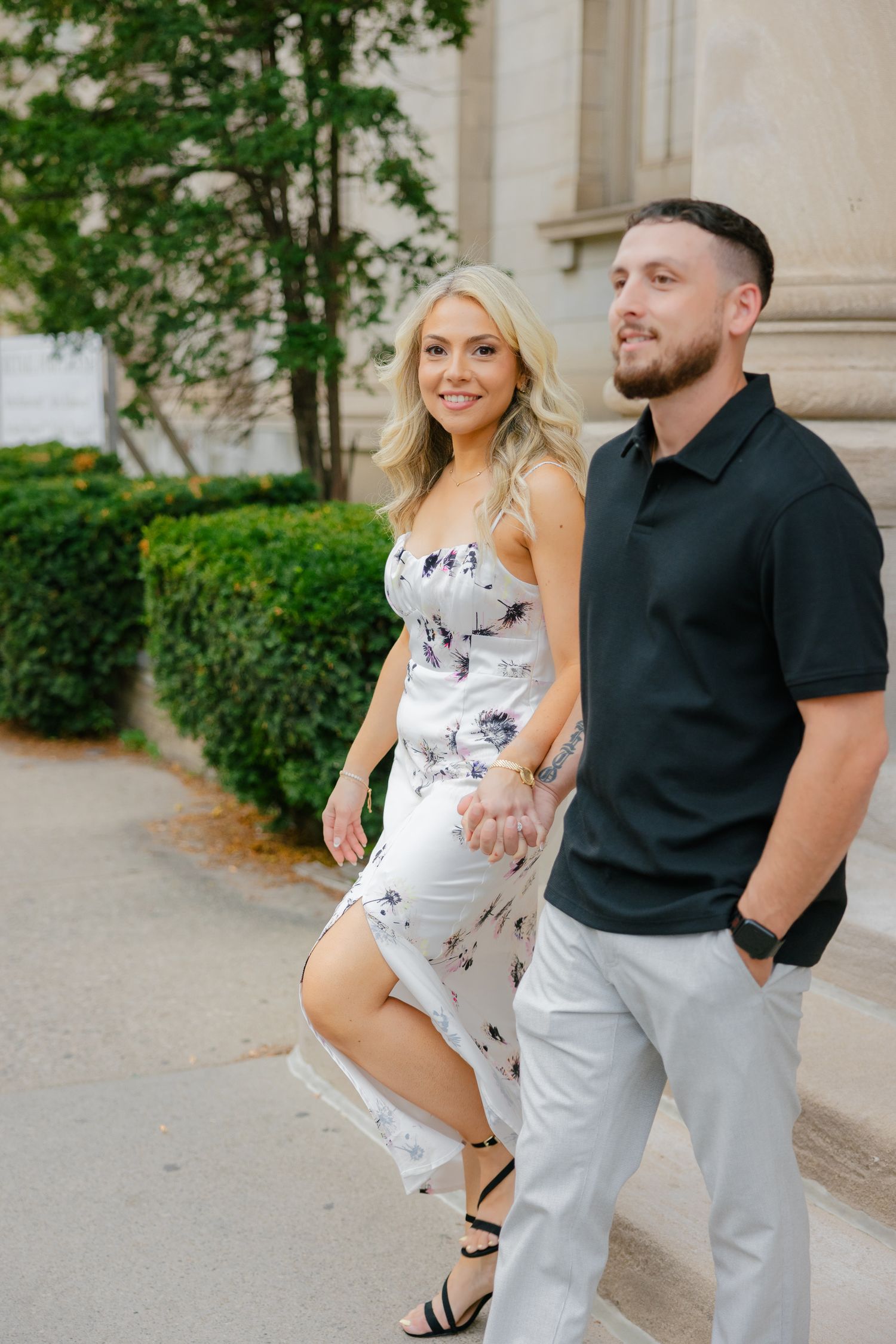 A couple poses against the limestone facade of a classical building on a sunny day.