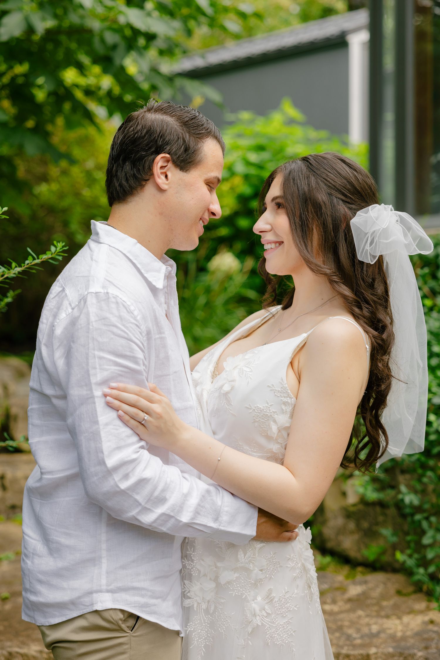 A romantic wedding portrait showing a couple in white embracing outdoors with lush greenery in the background.