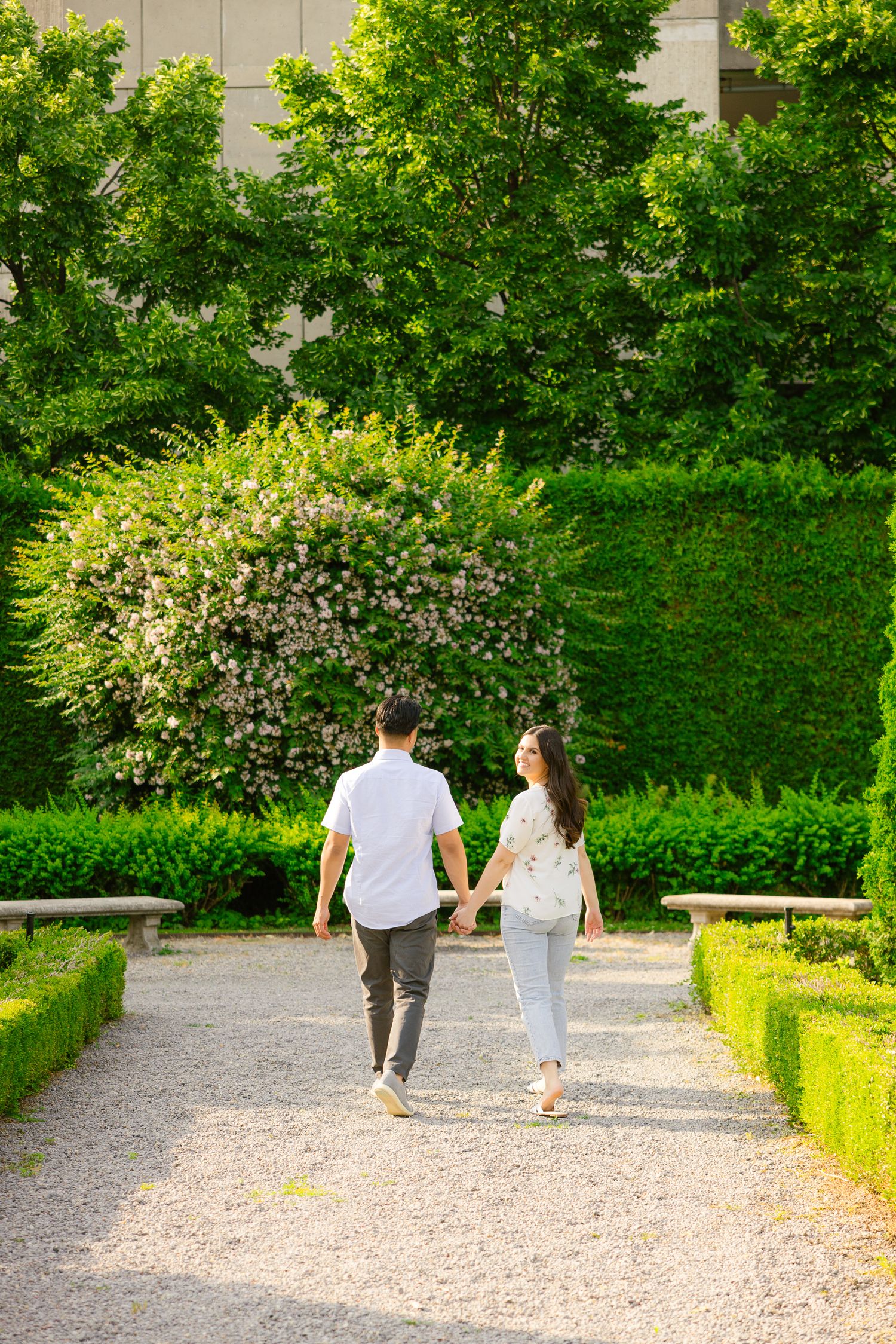 A couple walking away from camera down a formal garden path lined with trimmed hedges.