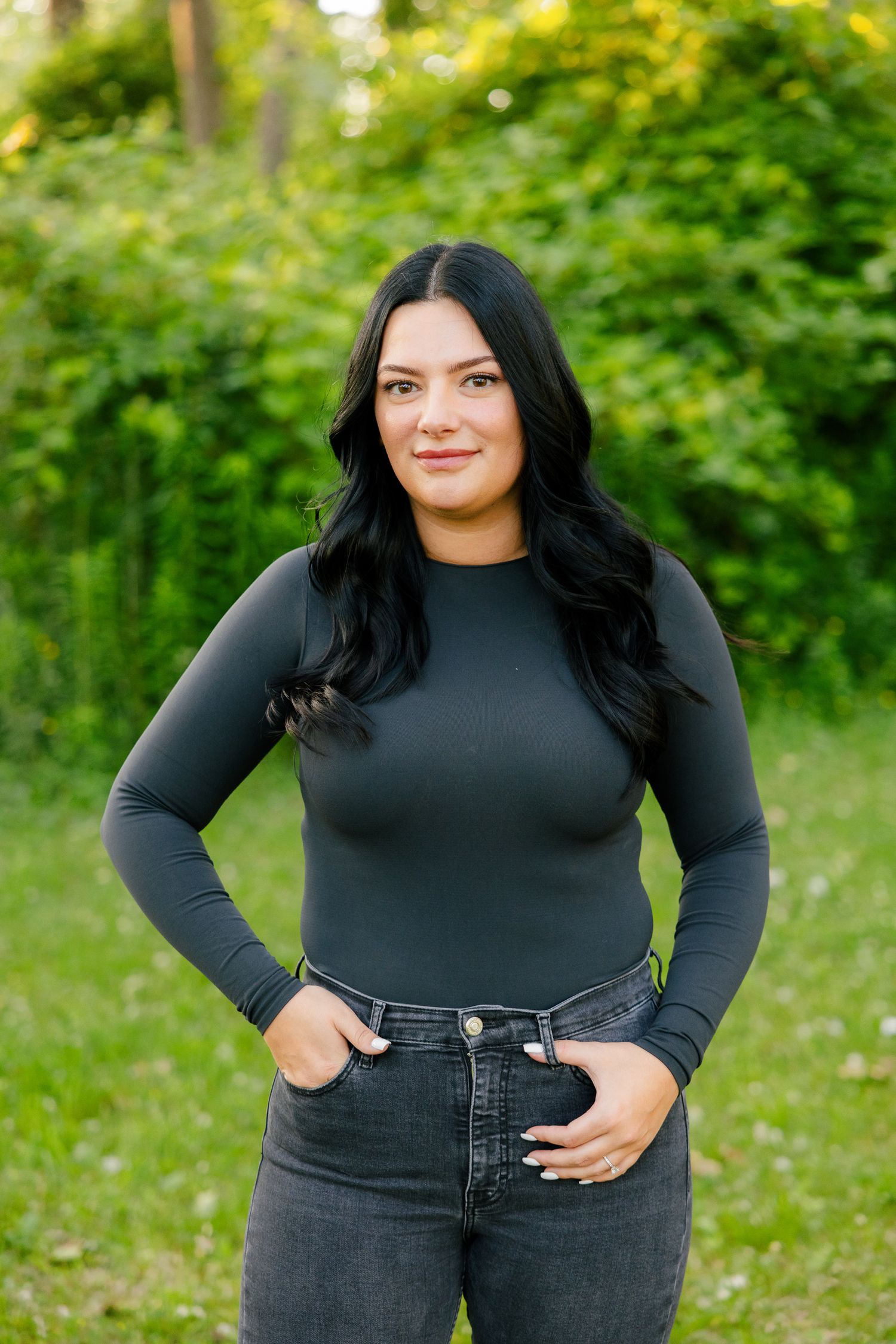 Woman in fitted black top and jeans poses confidently against lush green forest background.