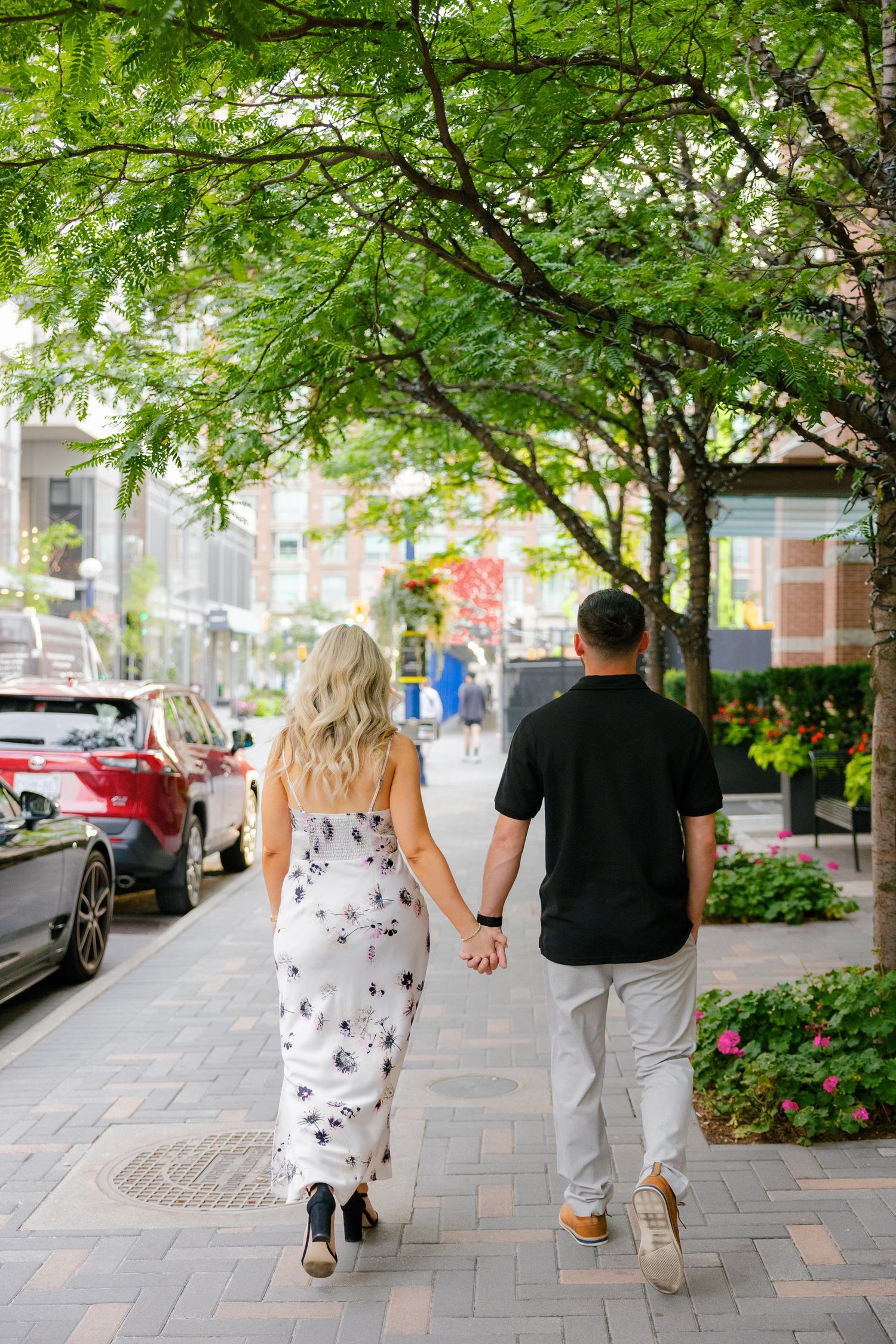 Couple strolling together on a city sidewalk under green leafy trees.