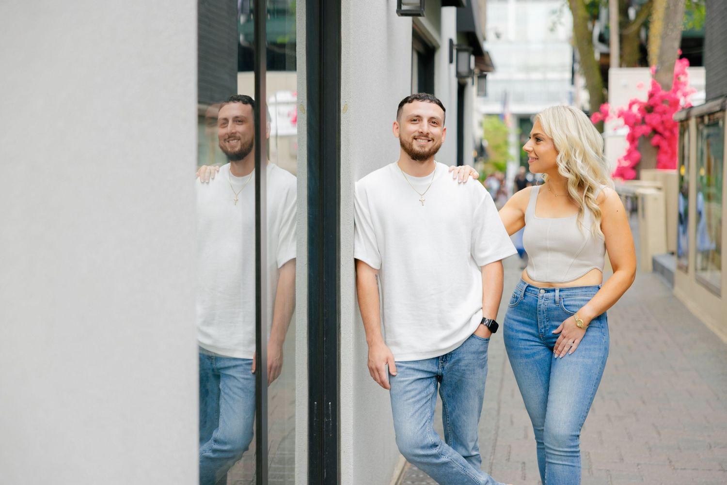 A couple in casual wear posing by reflective storefront windows on a city street.