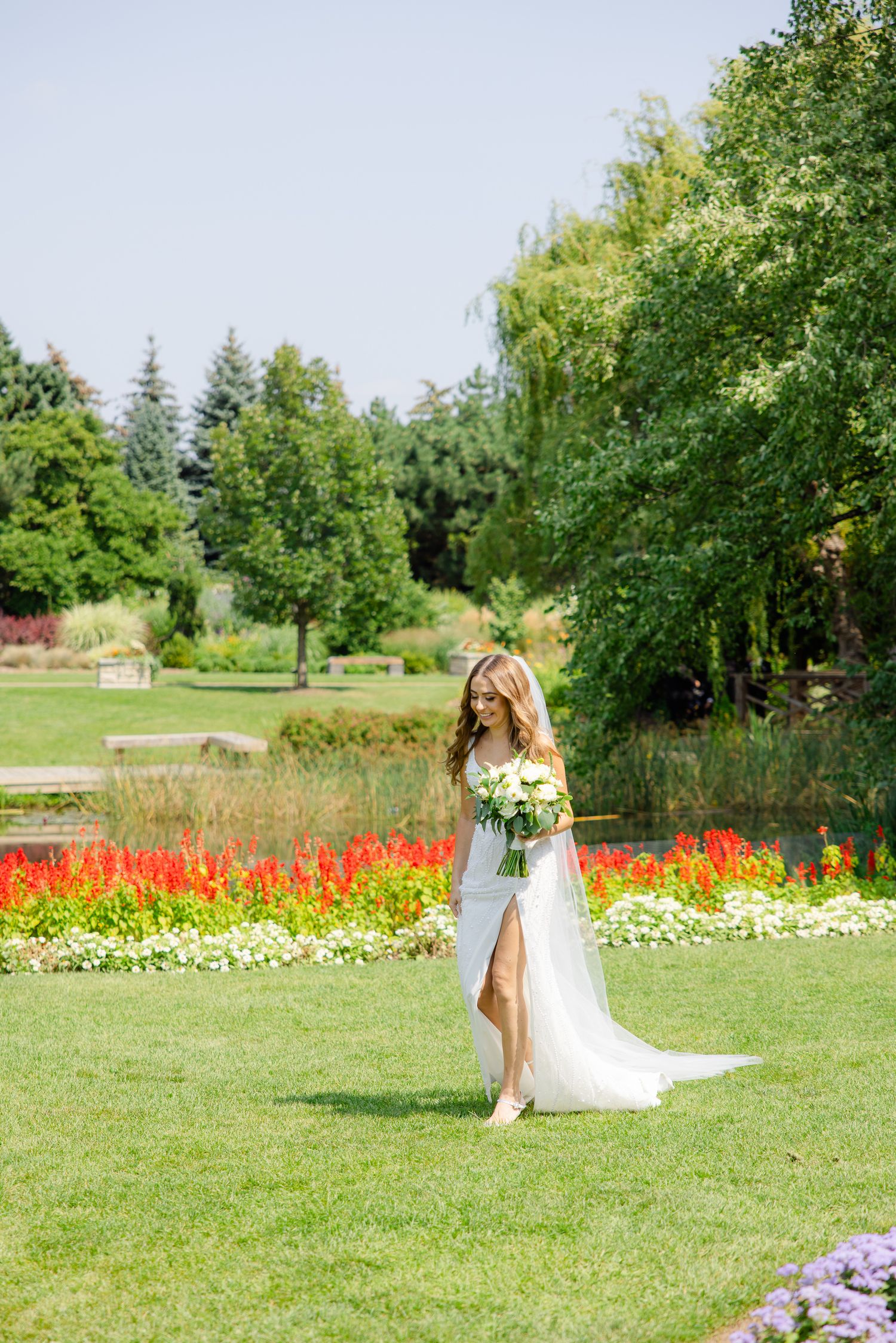 A bride in a flowing white dress poses in a garden with vibrant red flowers and lush greenery.