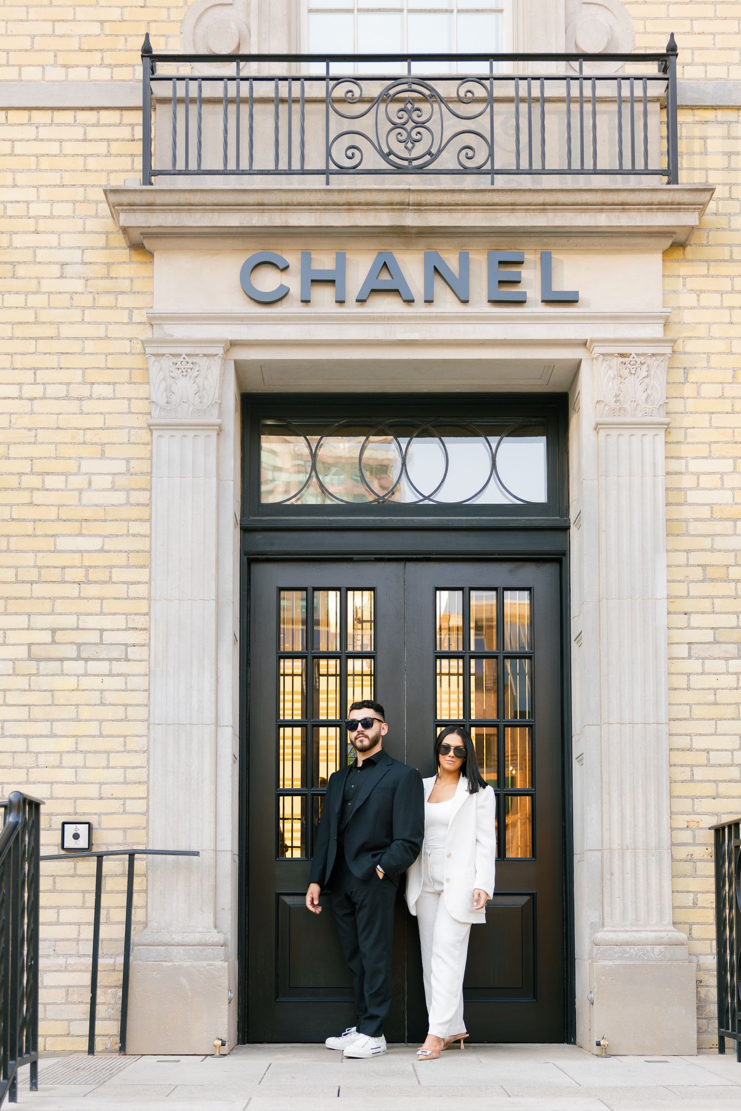 Couple in black and white outfits pose elegantly in front of Chanel store entrance.