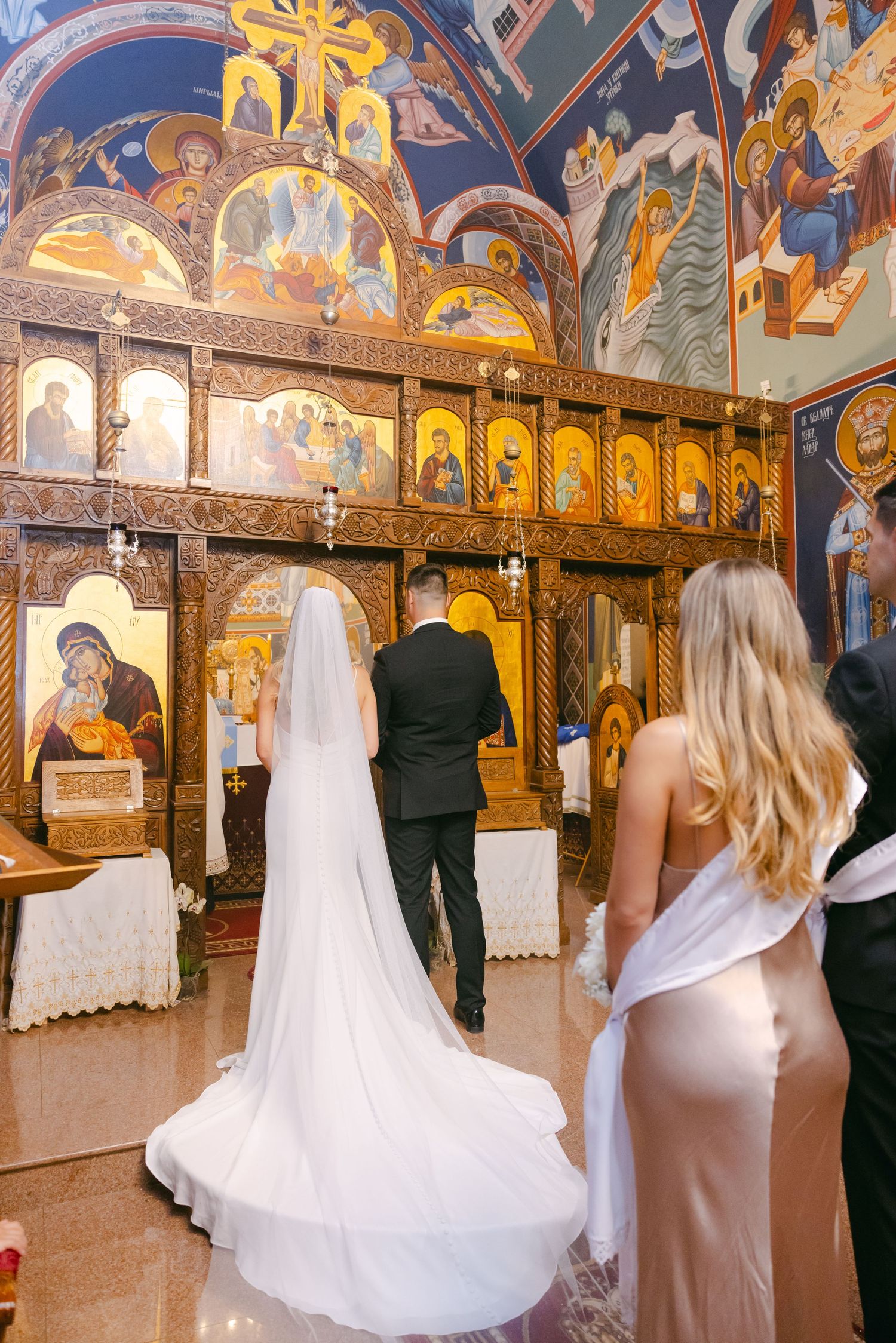 Wedding ceremony in progress inside an Orthodox church with beautiful iconostasis and religious murals.