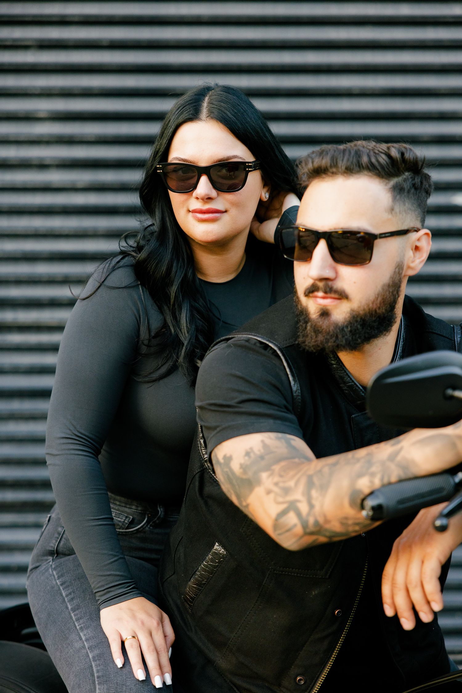 Couple wearing sunglasses sits on motorcycle together against modern metal background.