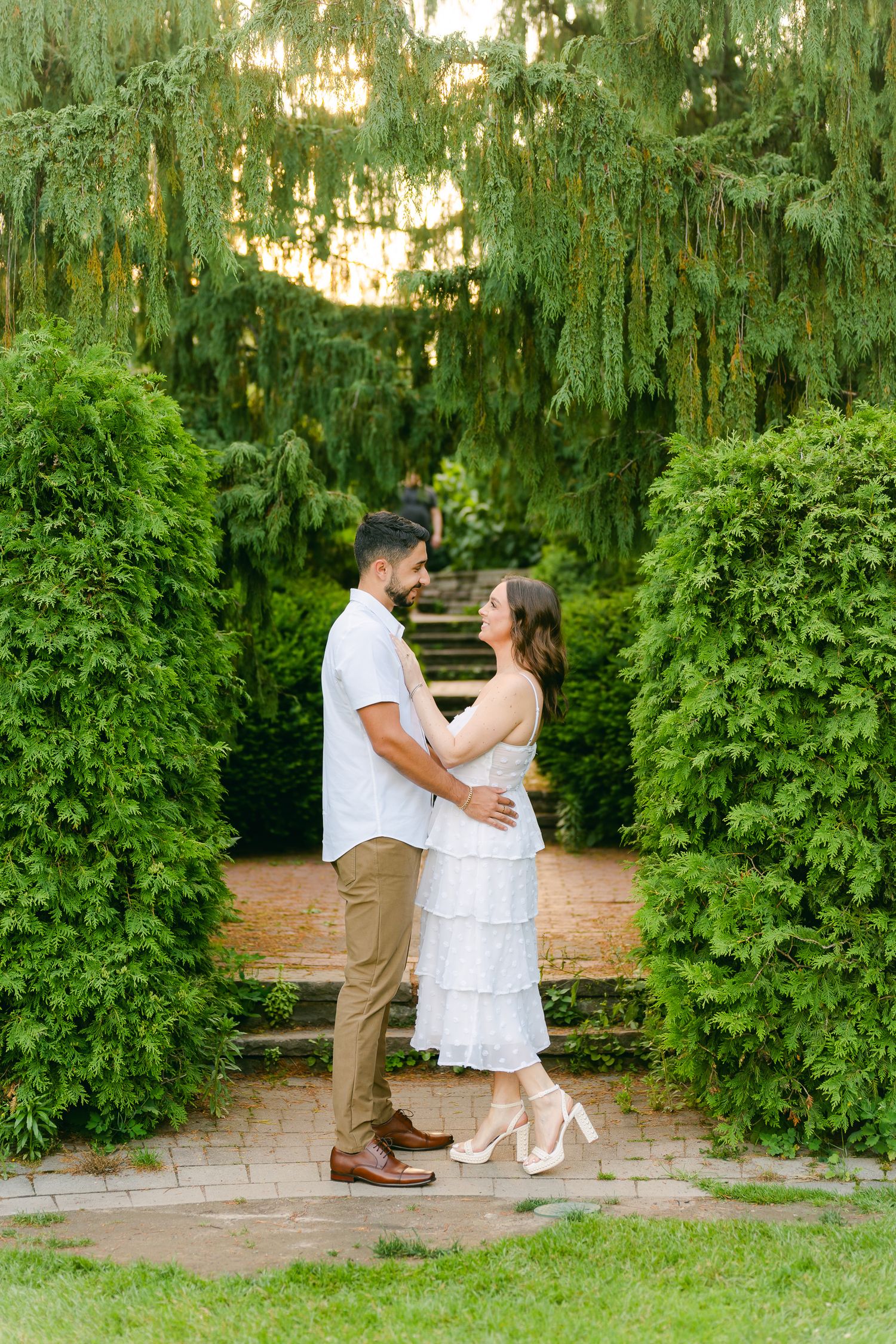 Couple in coordinated white and khaki outfits share an intimate moment on garden steps between manicured hedges.