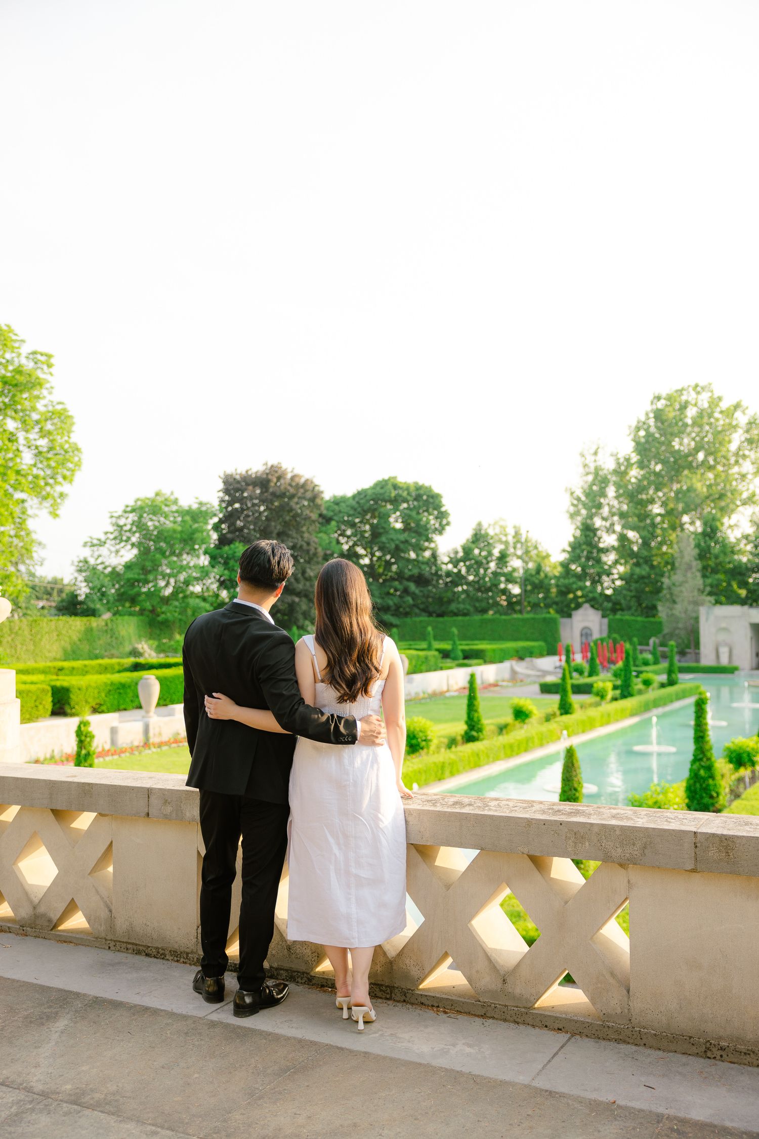 A couple overlooking beautiful formal gardens from a decorative stone balcony at sunset.