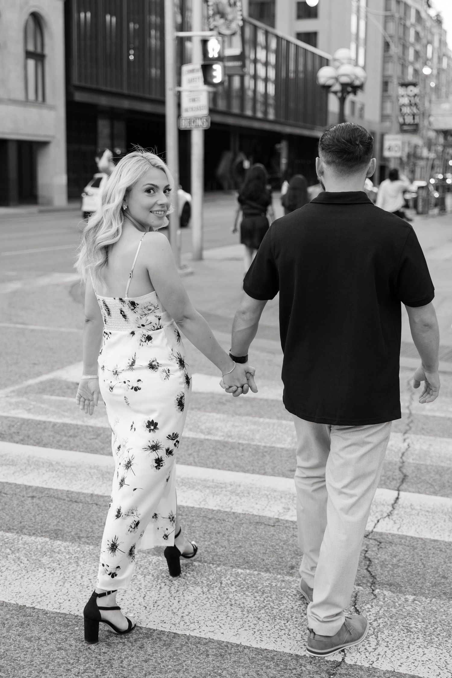 A couple holds hands while walking across a city crosswalk in black and white.