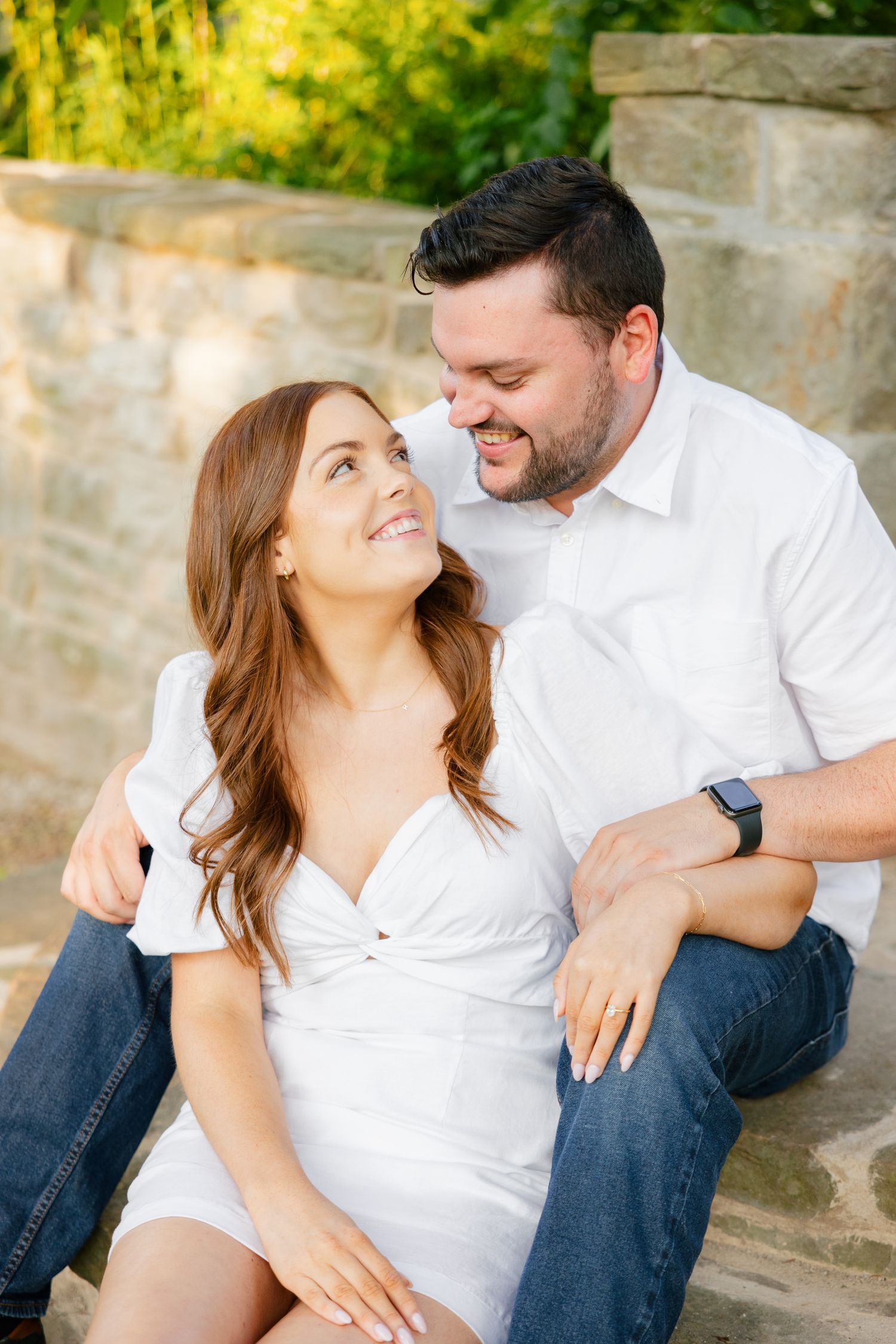 Couple in white clothing share an intimate moment while sitting on stone steps in a sunlit garden setting.