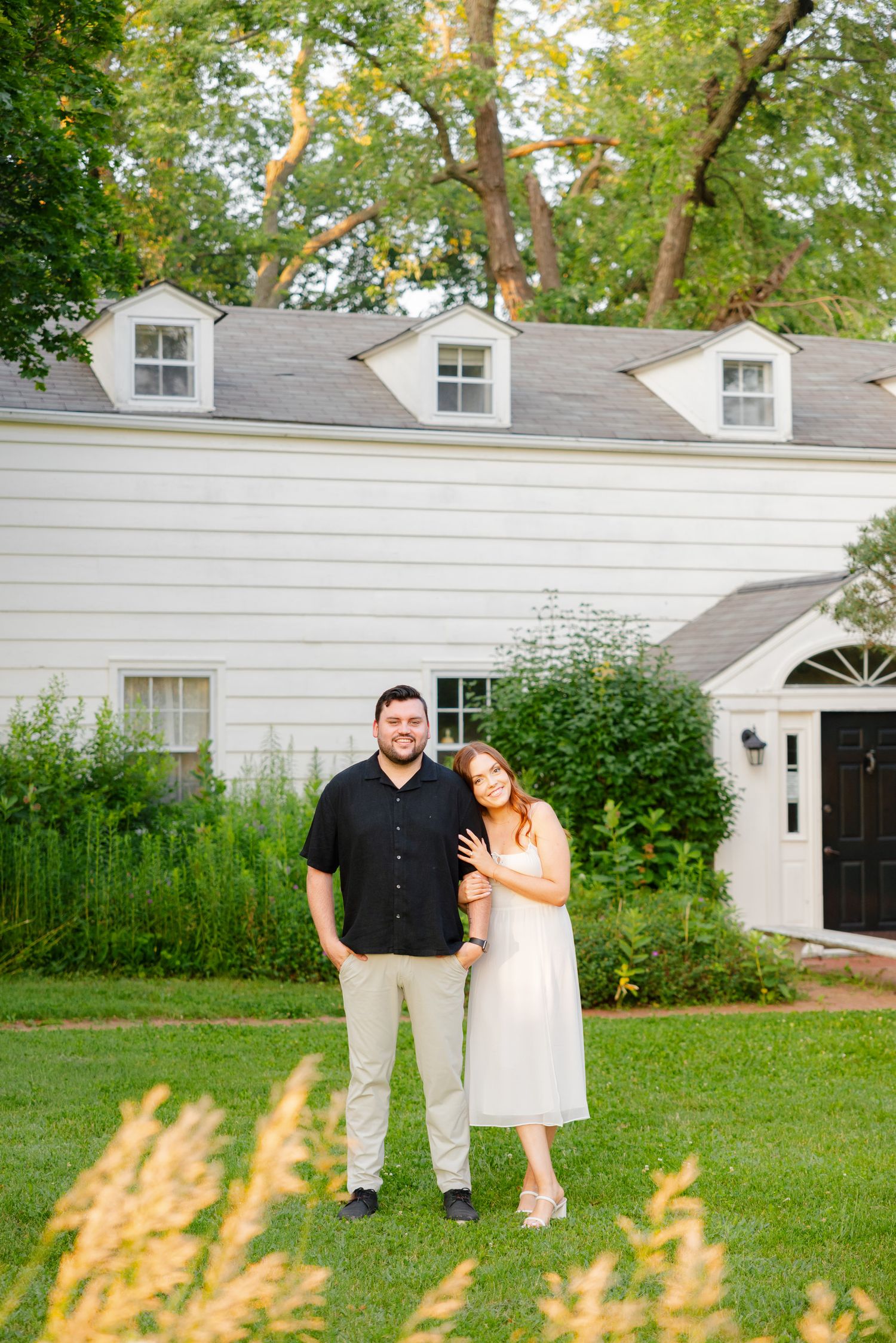 Couple posing on lawn in front of white colonial-style house with three dormer windows.