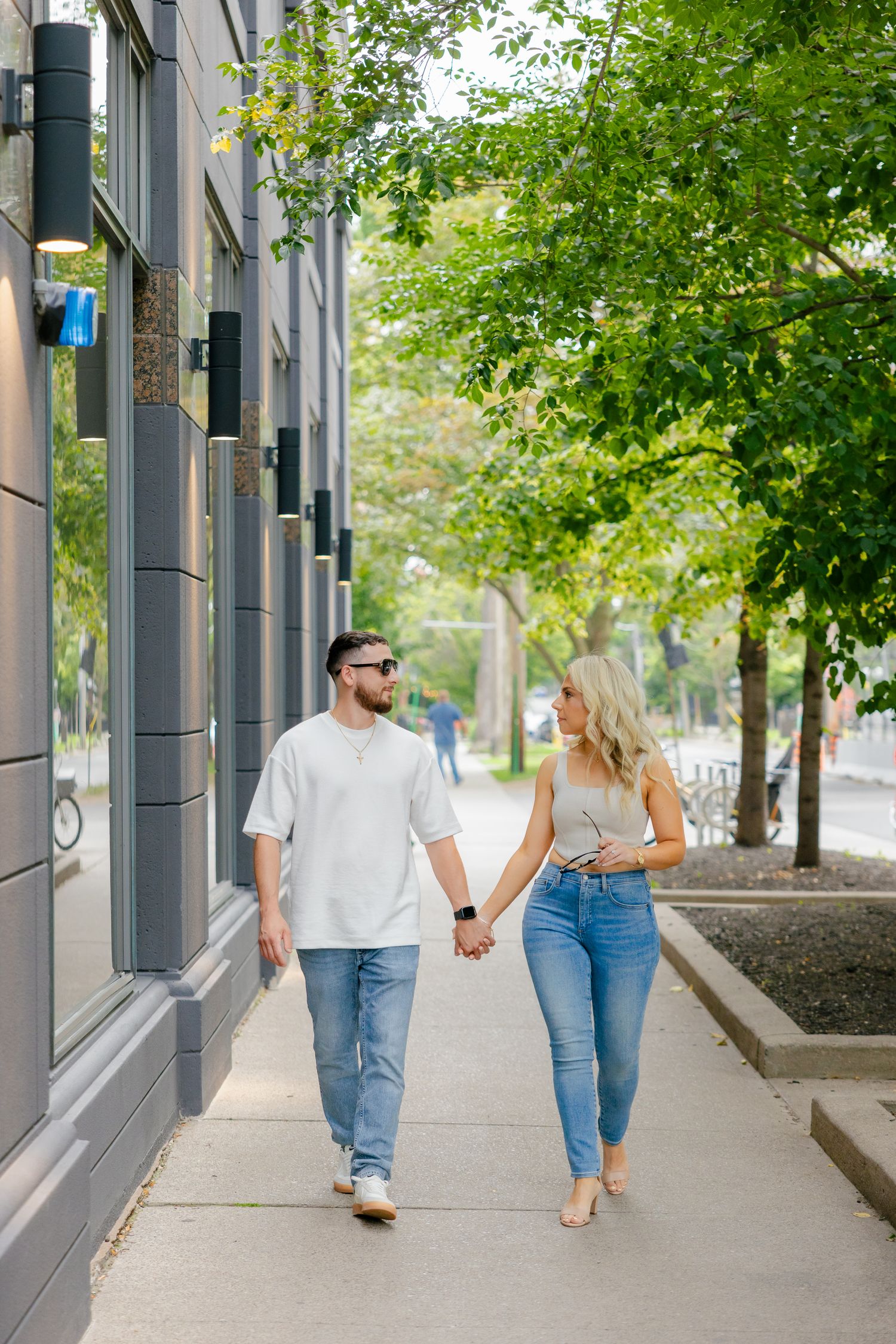 Couple in casual wear holding hands while walking along a city sidewalk lined with trees.