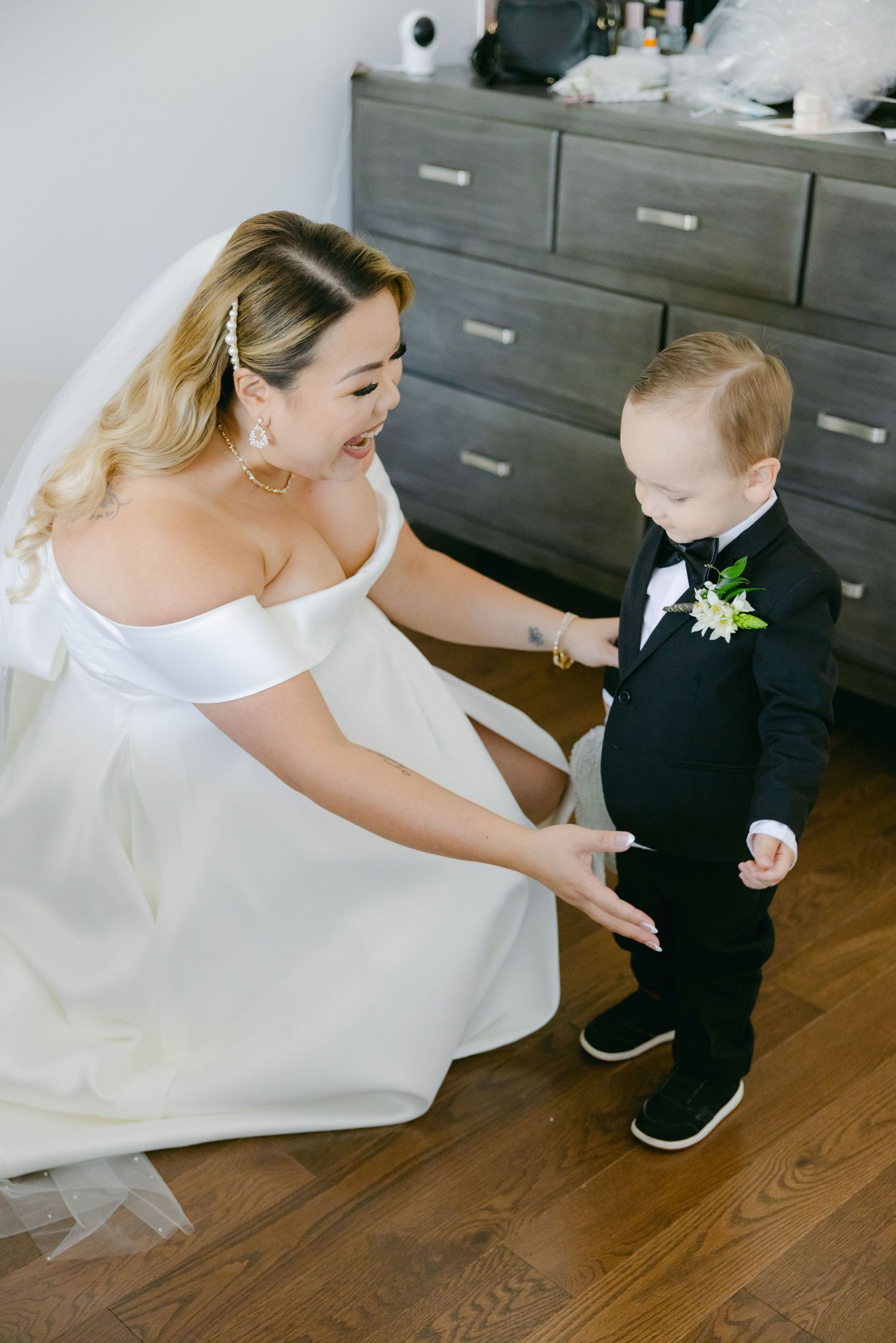 A bride in white kneels beside a young ring bearer wearing a tiny black tuxedo.