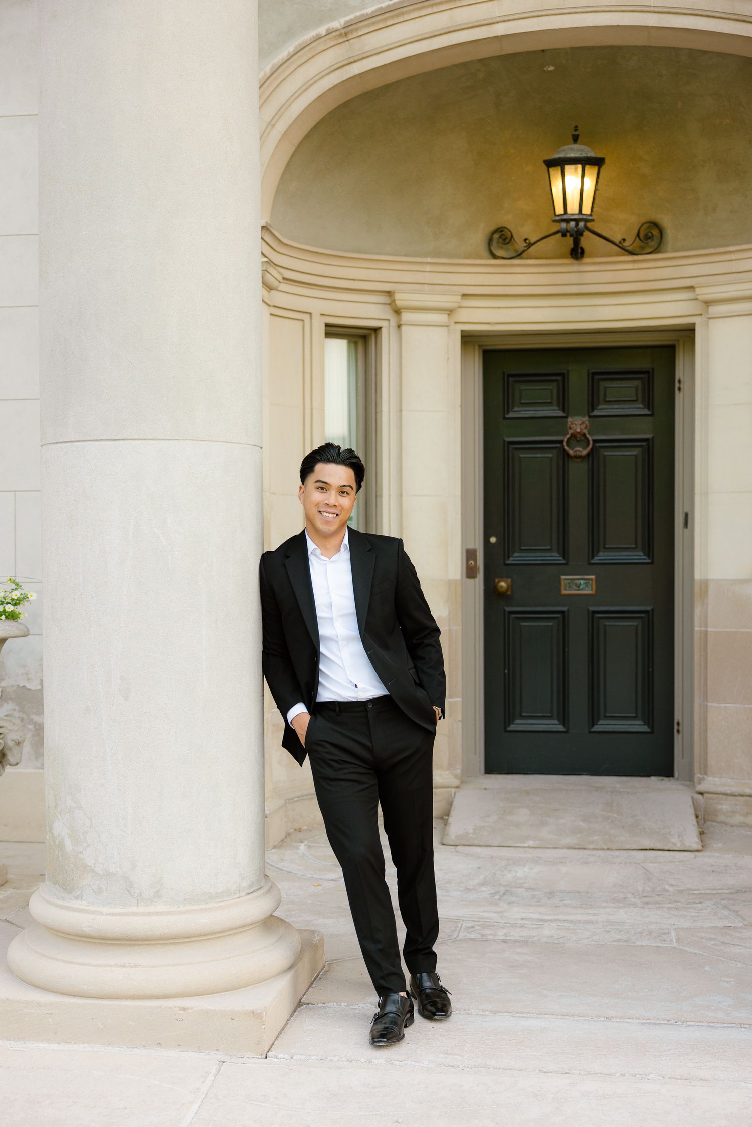 Man in black suit poses casually by elegant doorway with vintage lantern.