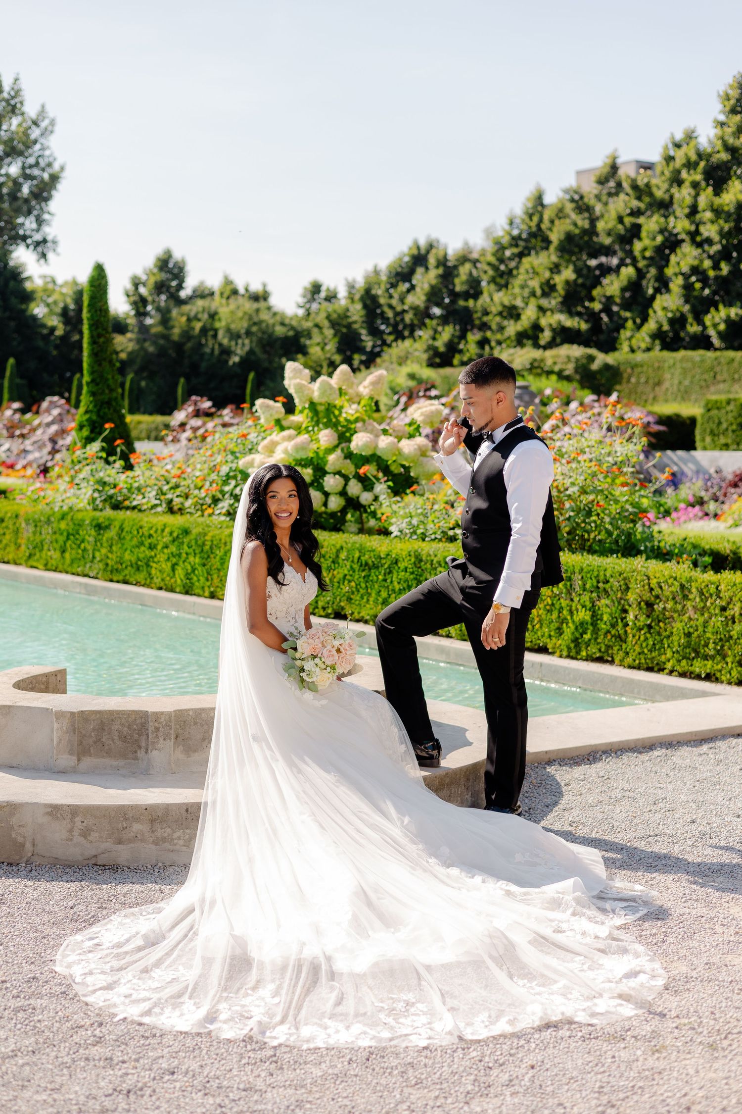 Wedding couple poses by a fountain in a formal garden with the bride's flowing train on display.