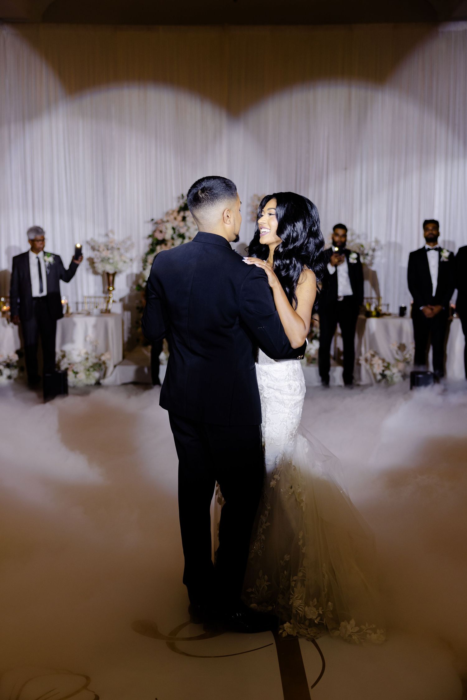 A couple embraces during their wedding dance surrounded by floral arrangements and white decor.