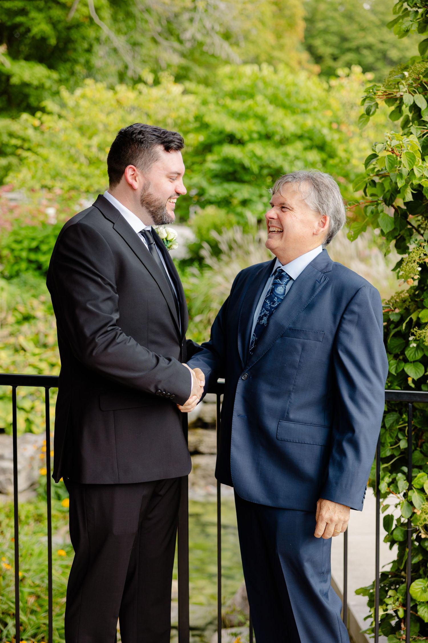 Two people in suits share conversation on wooden deck with garden background.