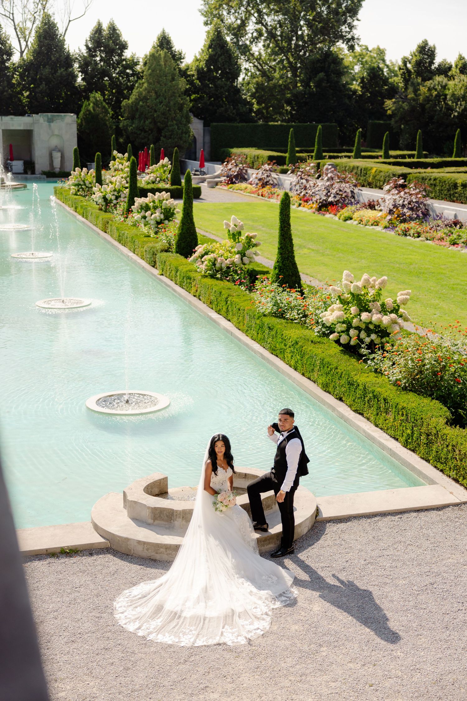 A couple in wedding attire photographed by a long ornamental pool with water features and landscaped gardens.