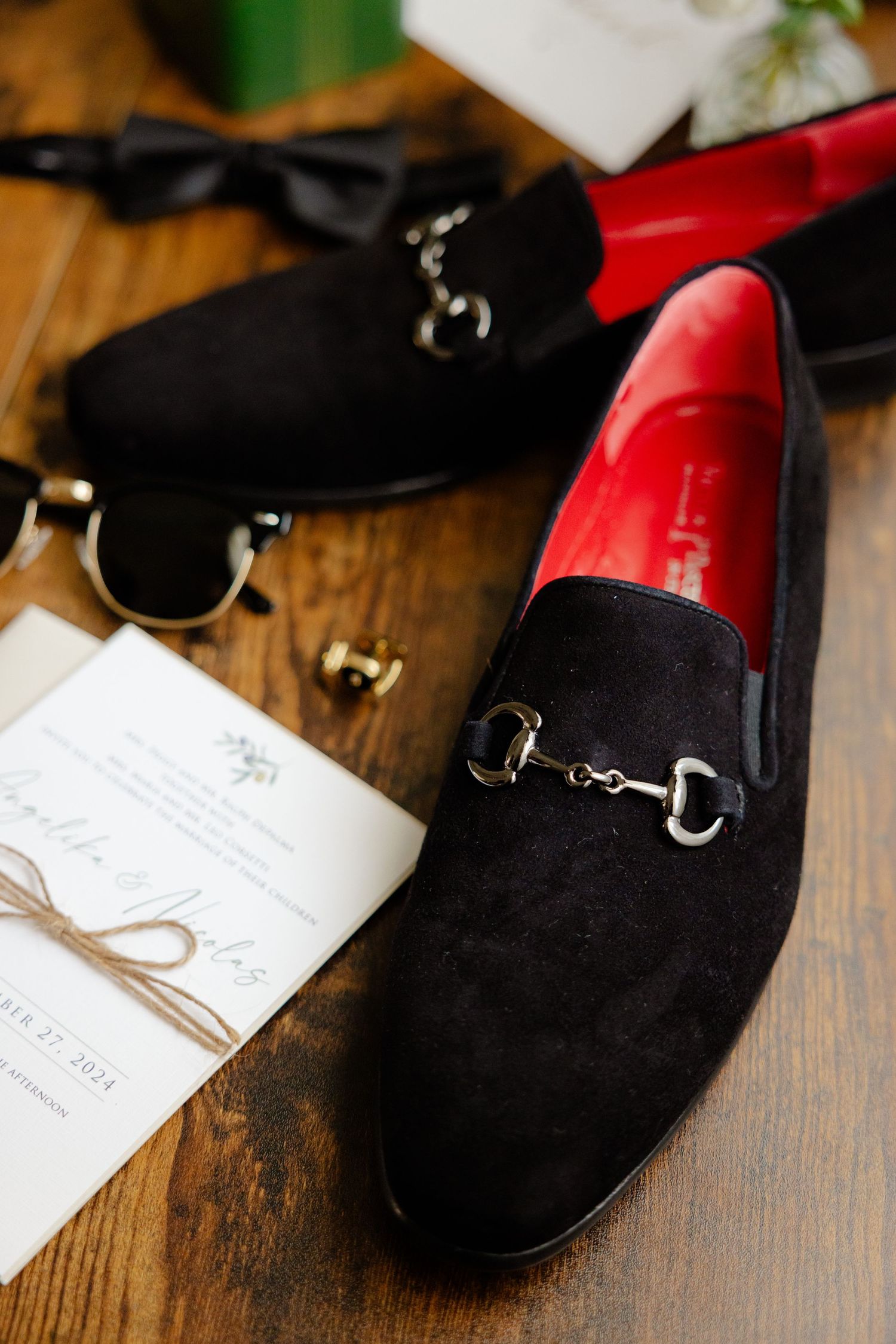 Black velvet loafers with metal hardware beside wedding invitation on wooden surface.