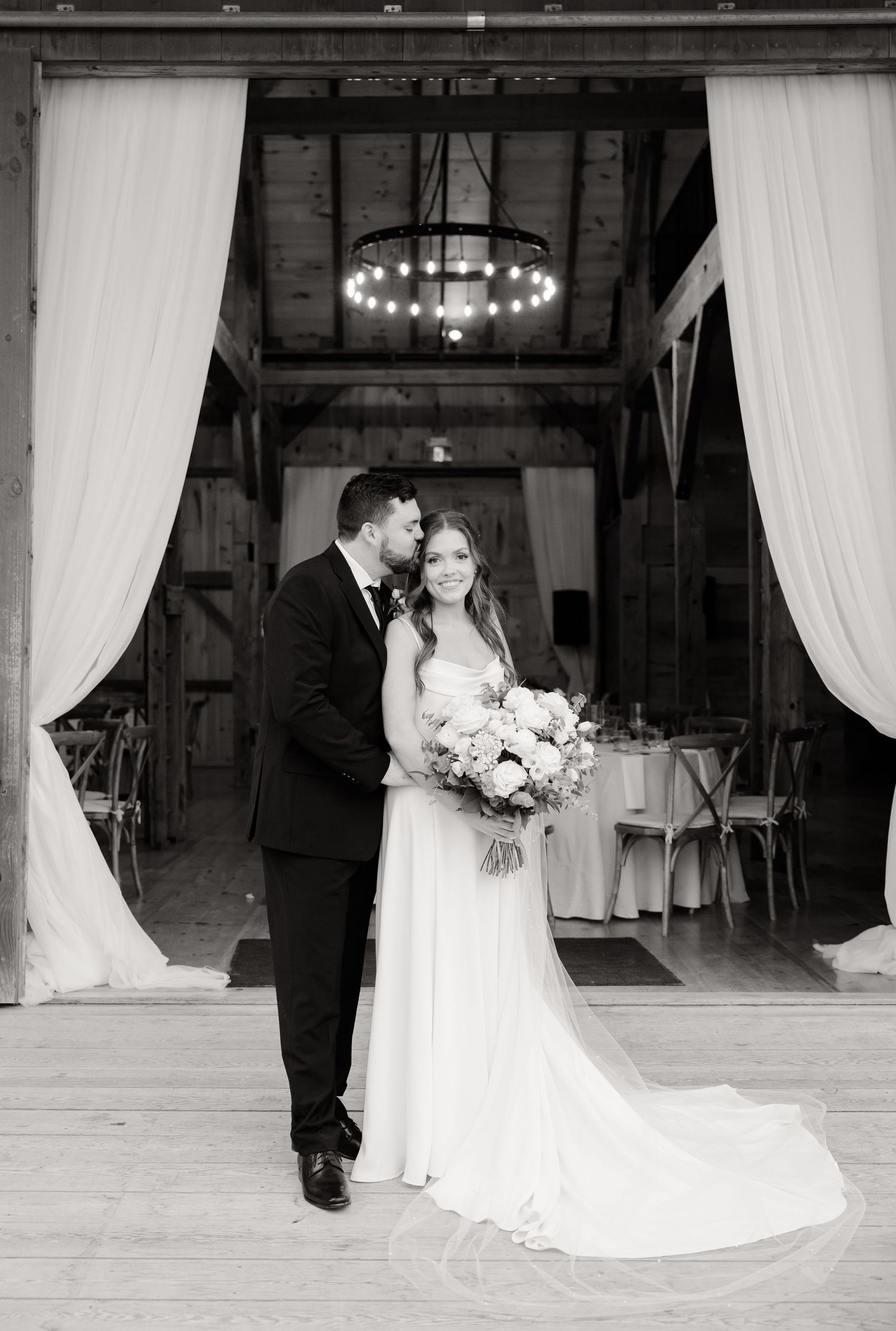 Black and white portrait of couple at rustic barn wedding venue entrance.