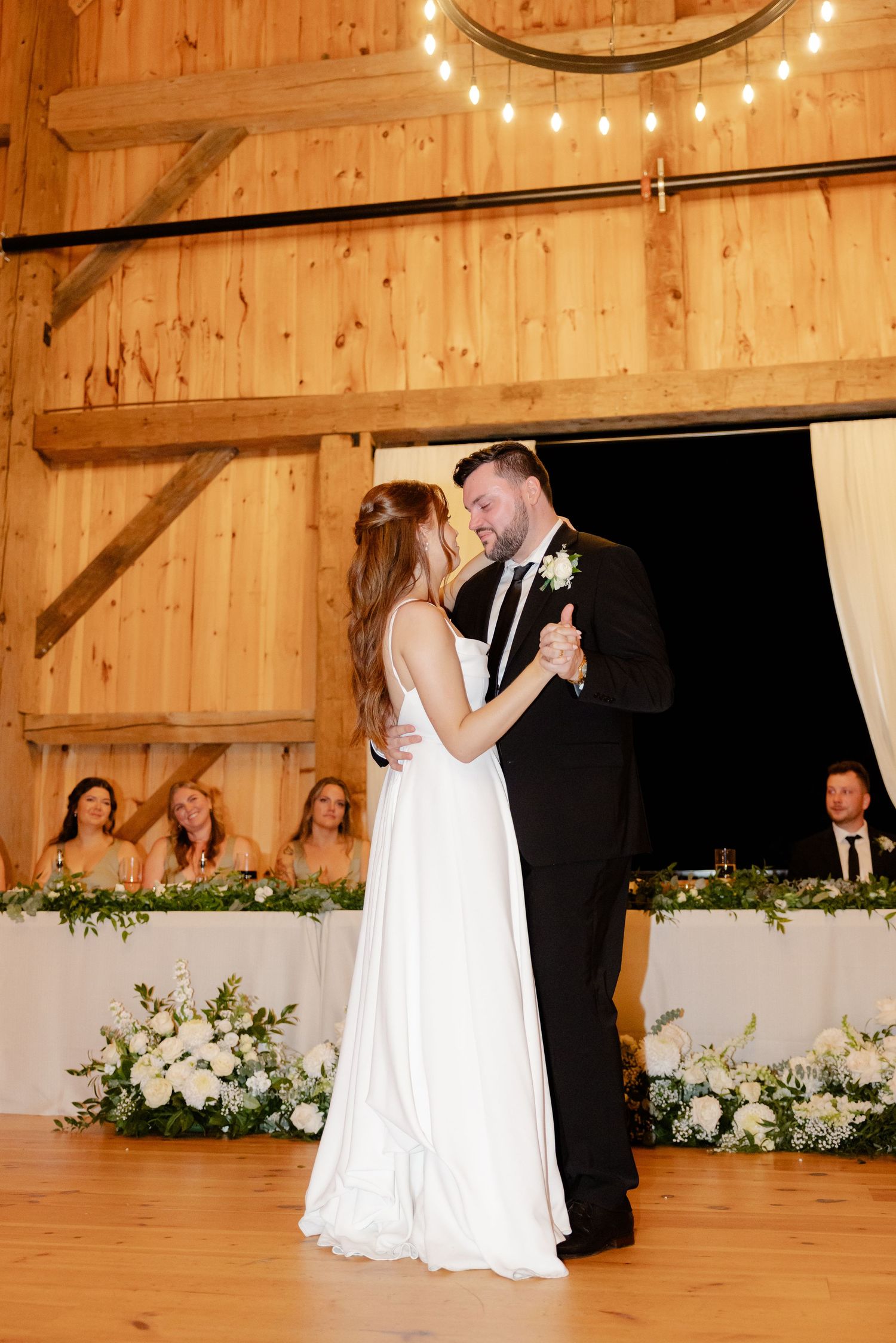 A couple shares their first dance under string lights in a beautifully decorated barn venue.