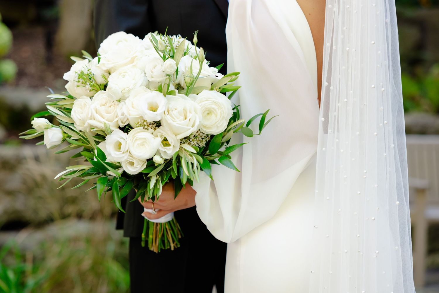 Close-up of white wedding bouquet with roses held against bride's elegant long-sleeve dress.