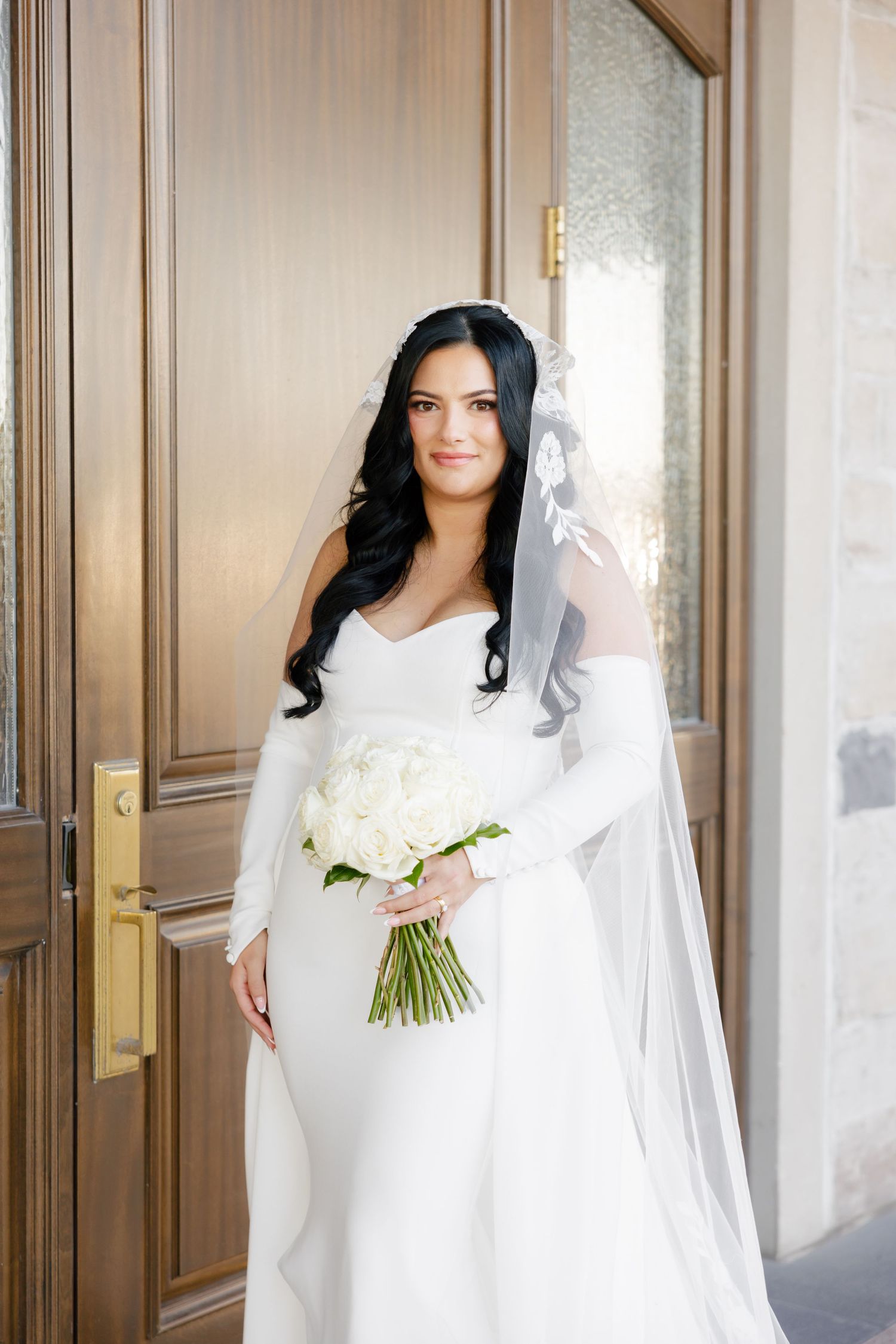 Bride in sweetheart neckline dress holds white bouquet while standing by wooden door.