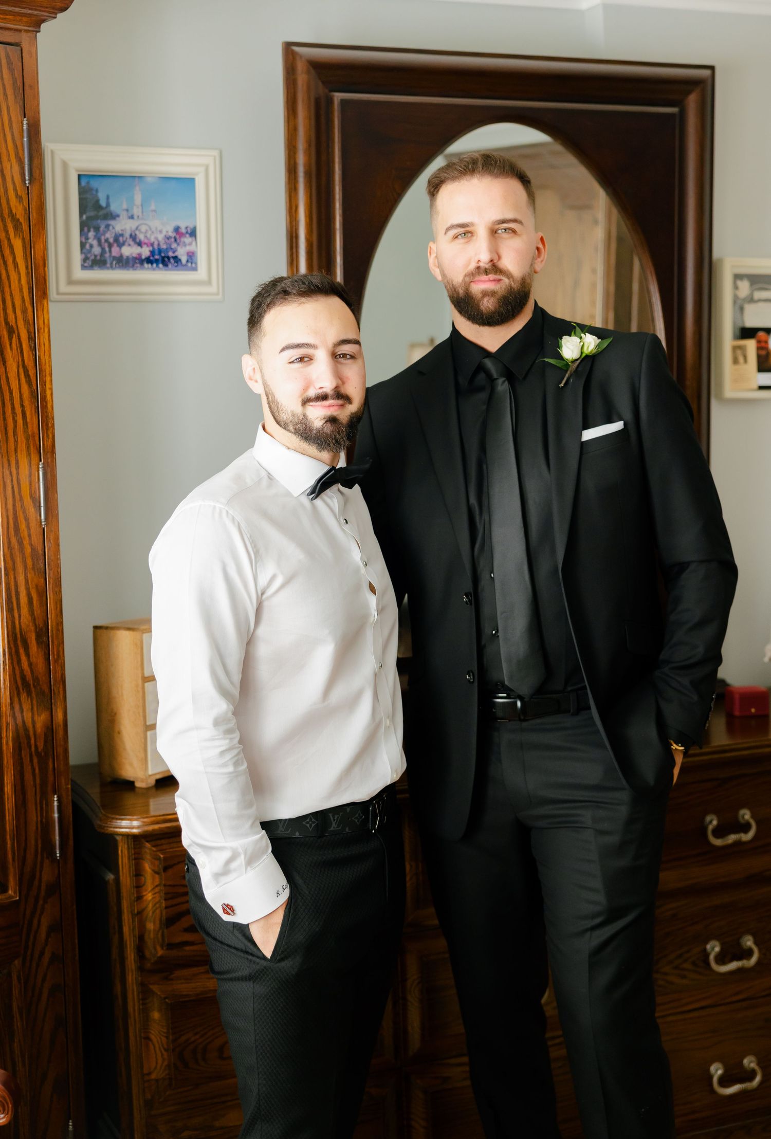 Two people in contrasting formal wear pose in front of an antique dresser with oval mirror.