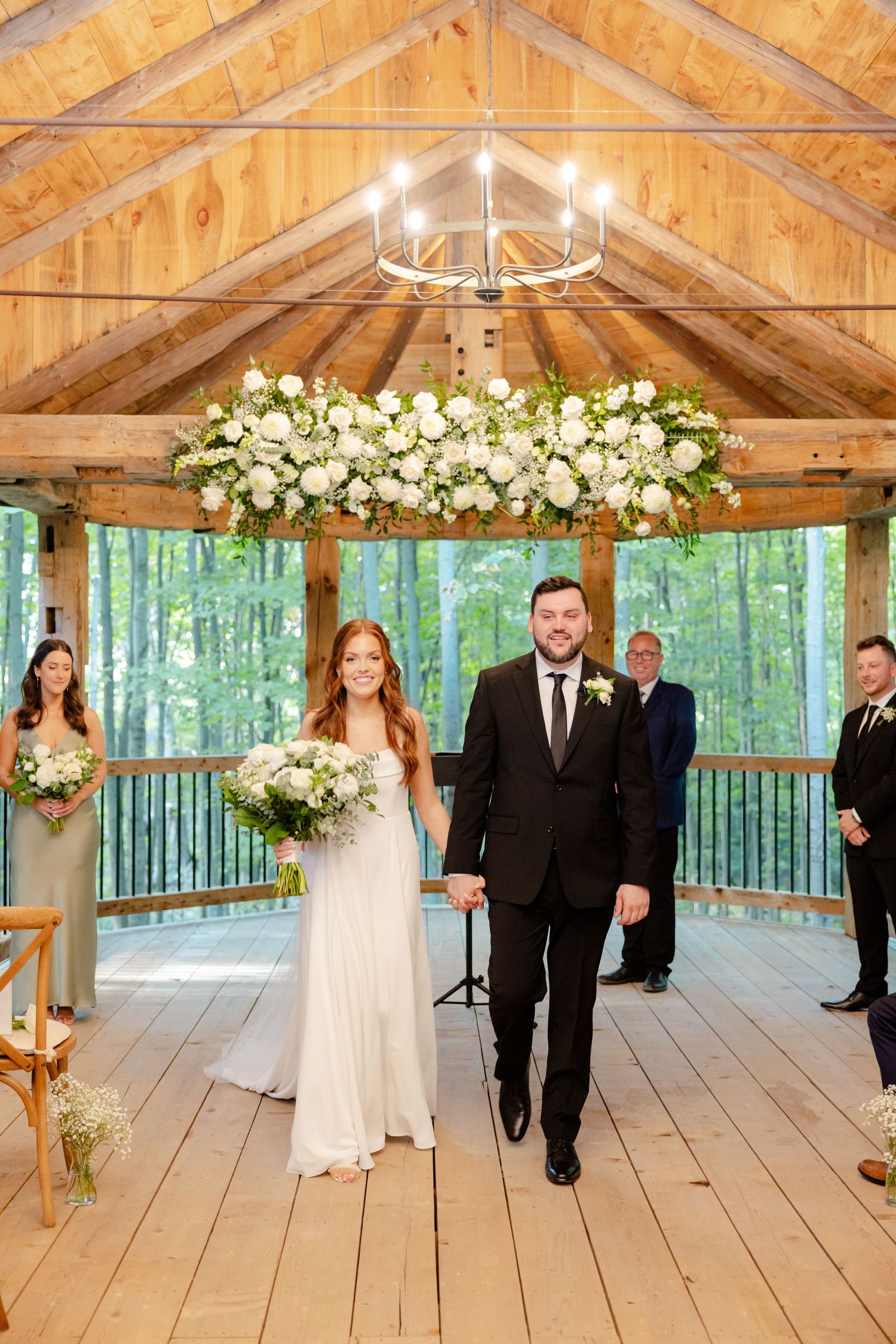 A wedding ceremony in progress inside a wooden pavilion decorated with white floral arrangements.